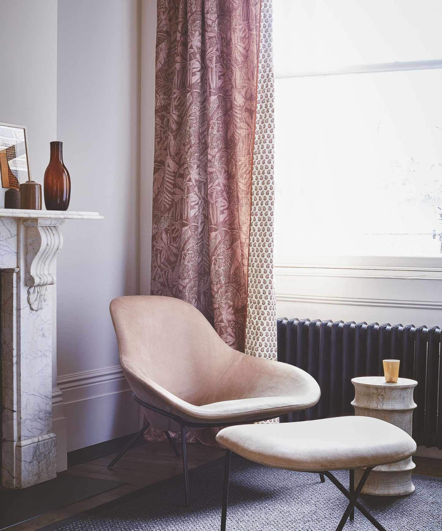 A neutral living room with pale pink walls and a marble mantelpiece. On top of the mantelpiece is a brown glass vase and framed picture. To the right is a peach velvet chair and matching footrest with a cylindrical marble coffee table with a glass on top. Behind is a black radiator underneath a large, bright window, with a pink and mauve patterned curtain open to the left-hand side.