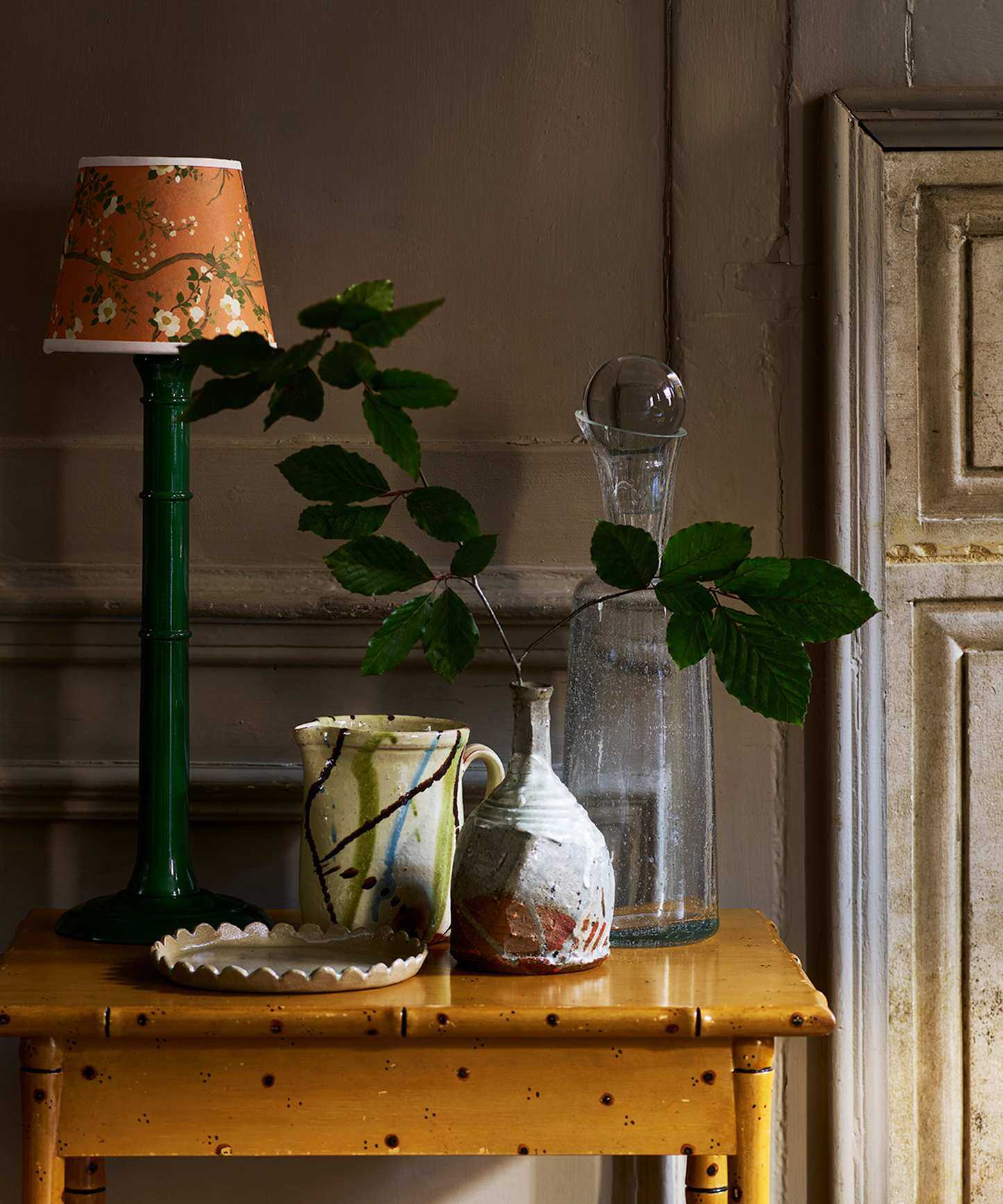 Ochre yellow painted side table with green table lamp, yellow decorative shade and ceramic vessels