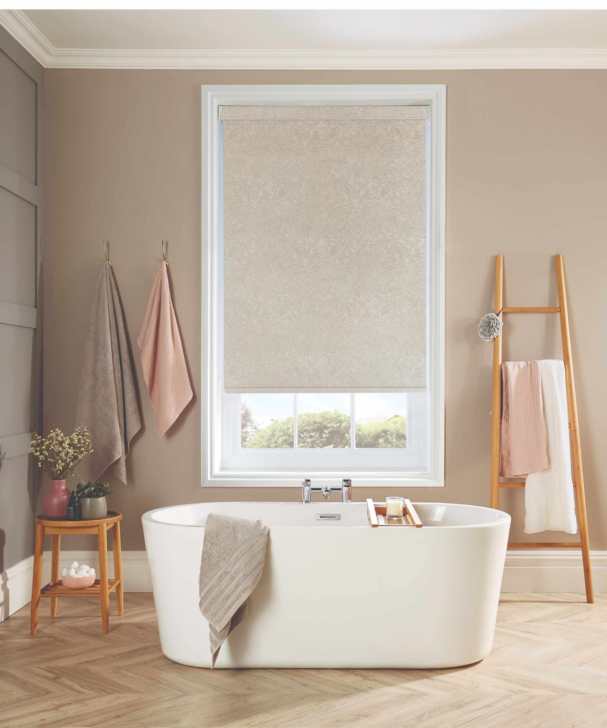 Freestanding white bath on wooden bathroom floor with neutral roller blind on central window in foregrounds. Peach, gray and taupe towels and wooden rails around the bathroom can be seen