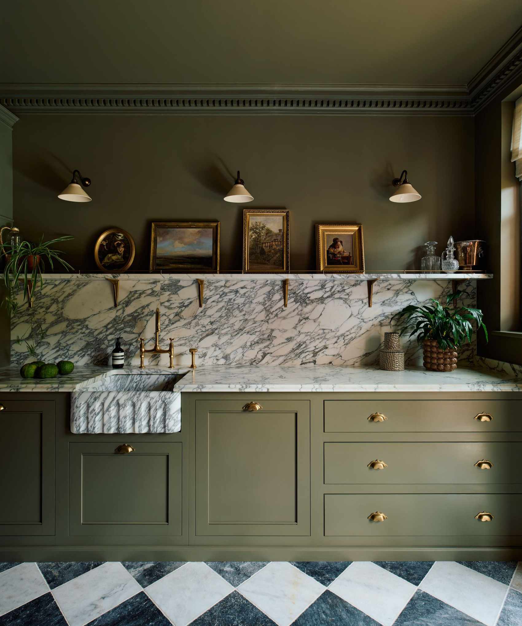 A classic kitchen with olive green cabinets and matching wall and ceiling, marble countertops and backsplash, a gallery shelf with antique picture frames, and sconce lighting on the wall.