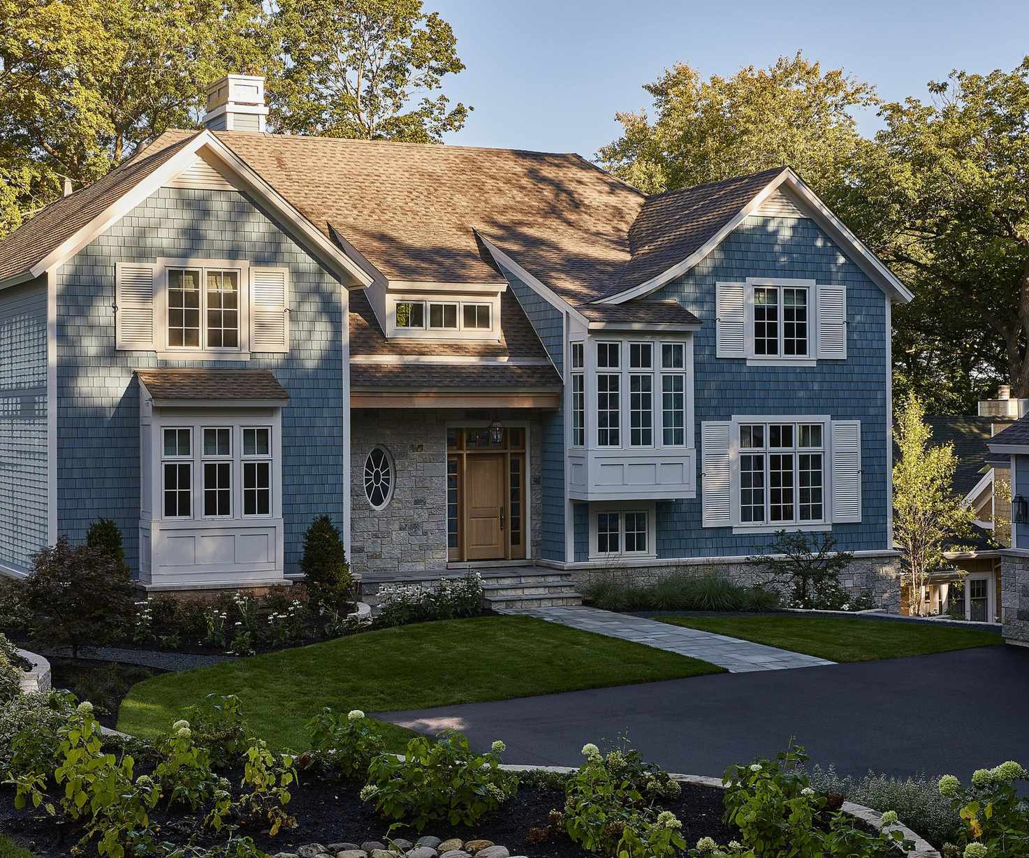 A home exterior with muted blue brickwork, white trim and window shutters, and a wooden front door. A front garden with lawn and plants, and a paved path.