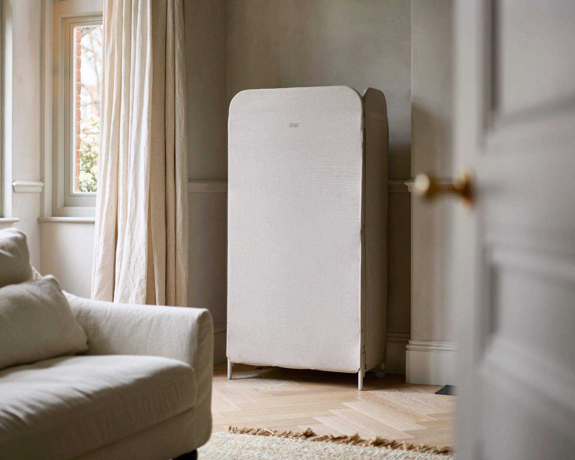 The Eclipse&trade; 3-tier Ecru Clothes Drying Rack With Screen turned away to conceal laundry in a large, bright living room
