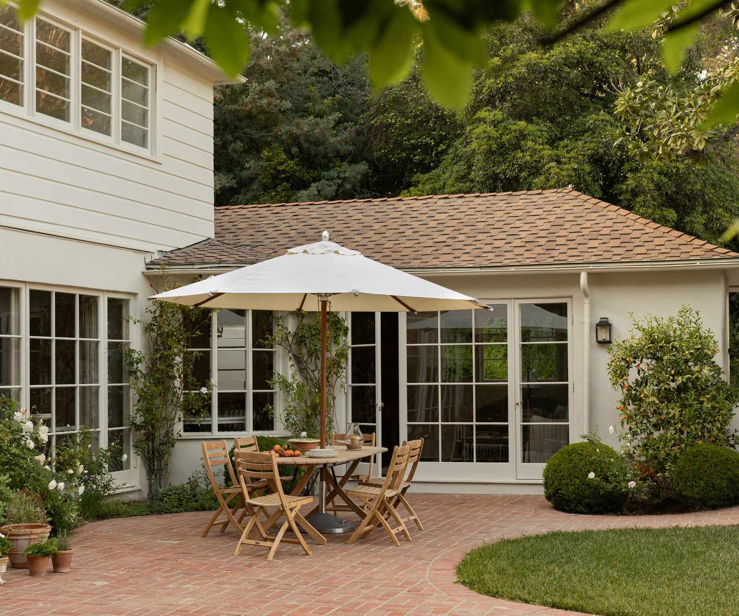 A home exterior painted warm white with a red brick roof, a patio area with a wooden table and chairs with a cream parasol, and planted shrubs.