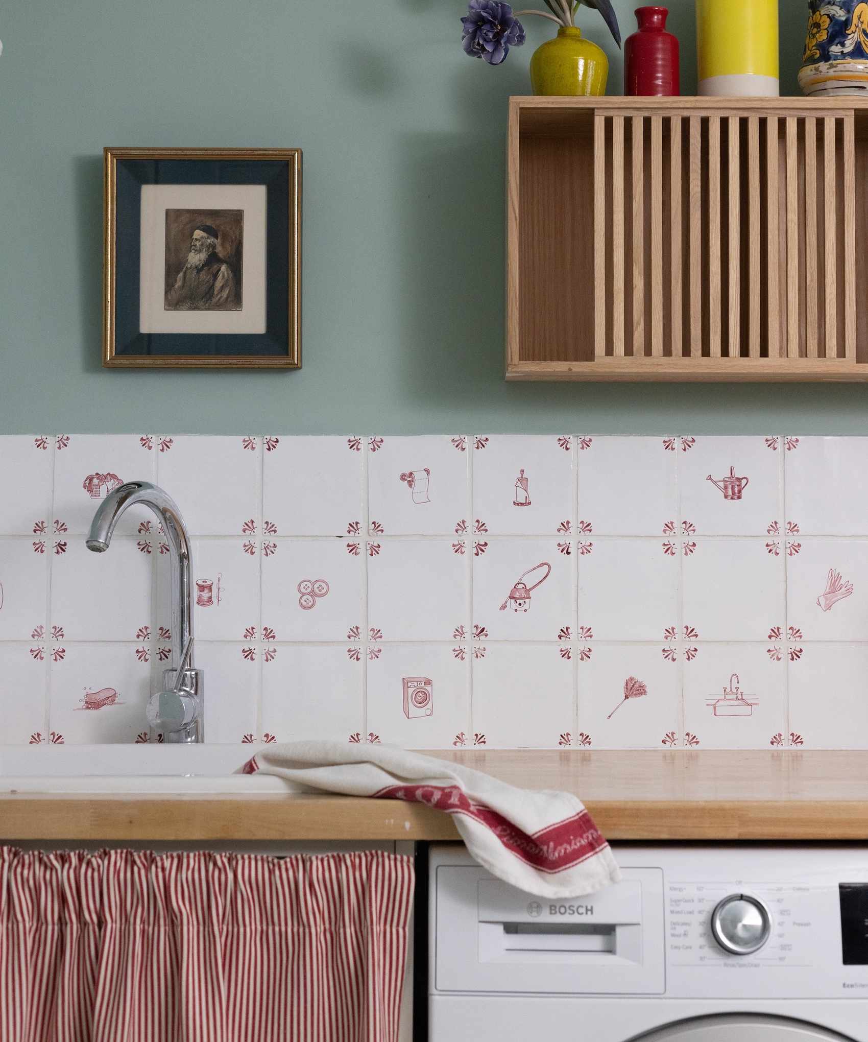 Quaint laundry room with whit and red patterned tile back splash, red striped curtain closure under sink and the top of a white washing machine under a wooden counter with ceramic white sink.