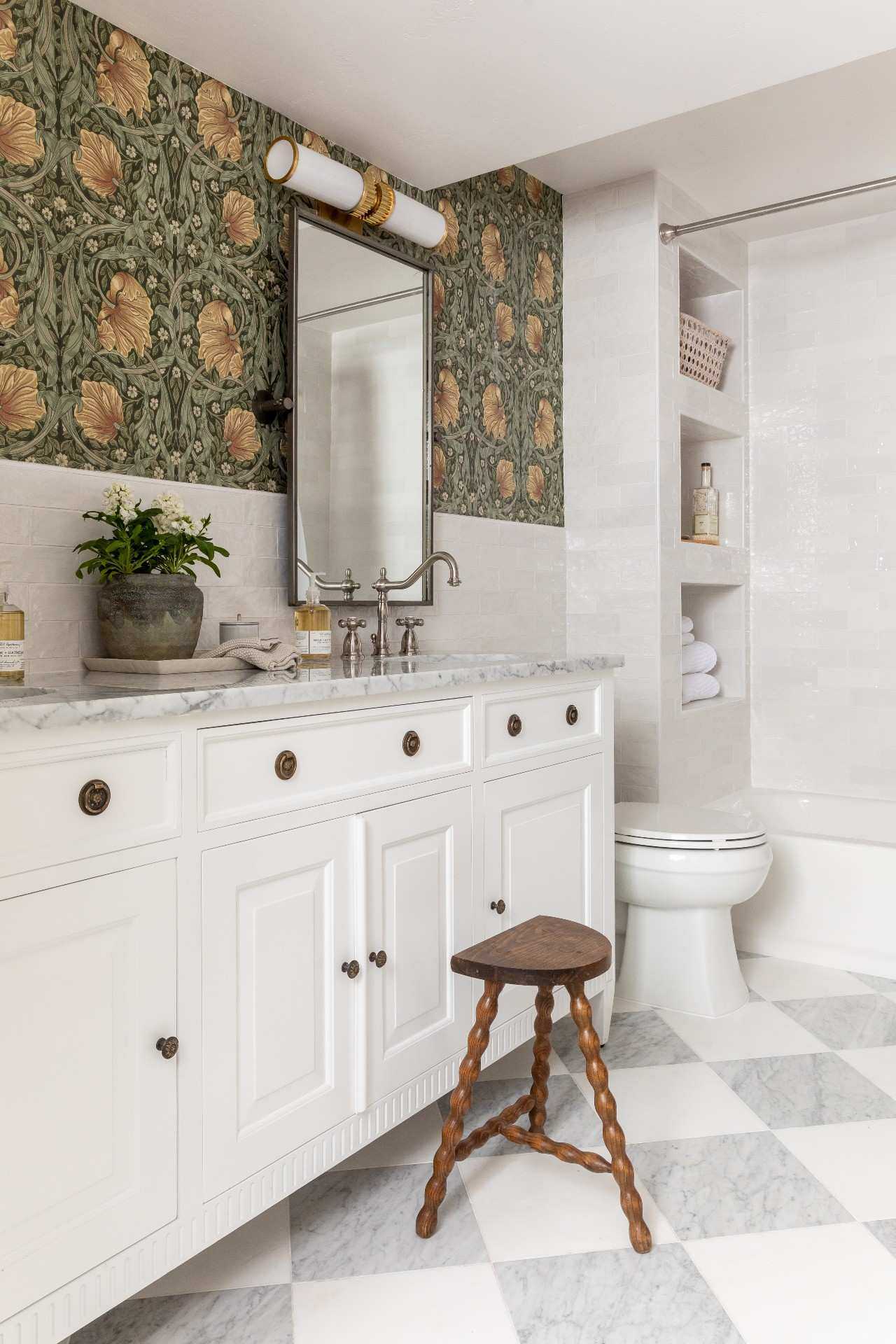 A basement bathroom, with white vanity cabinets and green floral patterned wallpaper on the walls. A white toilet and shower in the background, and a wooden stool in the foreground. A grey and white checkered floor.