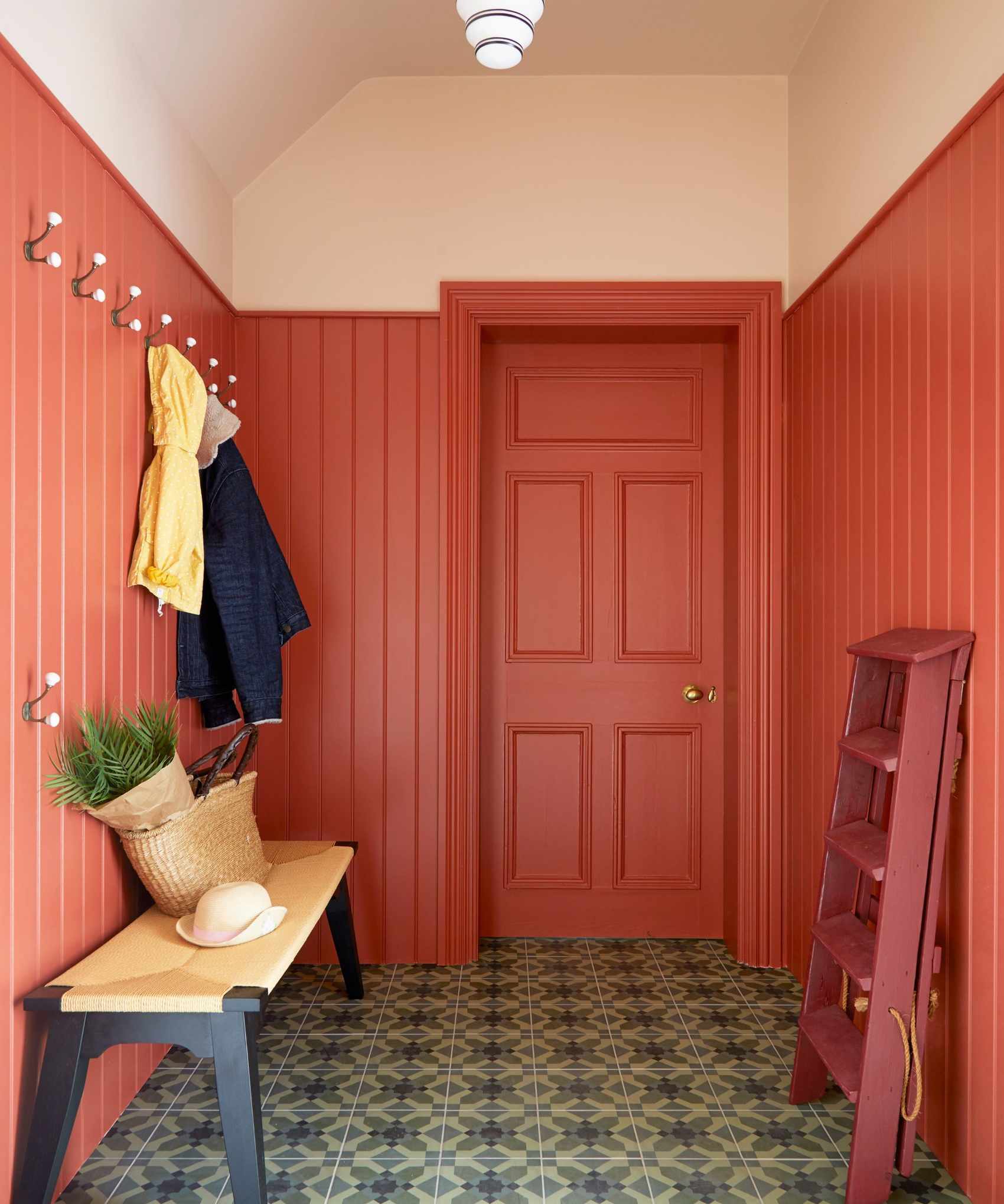 A boot room with panelled walls painted in a vivid coral paint. There are hooks on the wall with rain jackets and a bench with a bag and plant on it. On the other side of the room is a red ladder leaning against the wall.