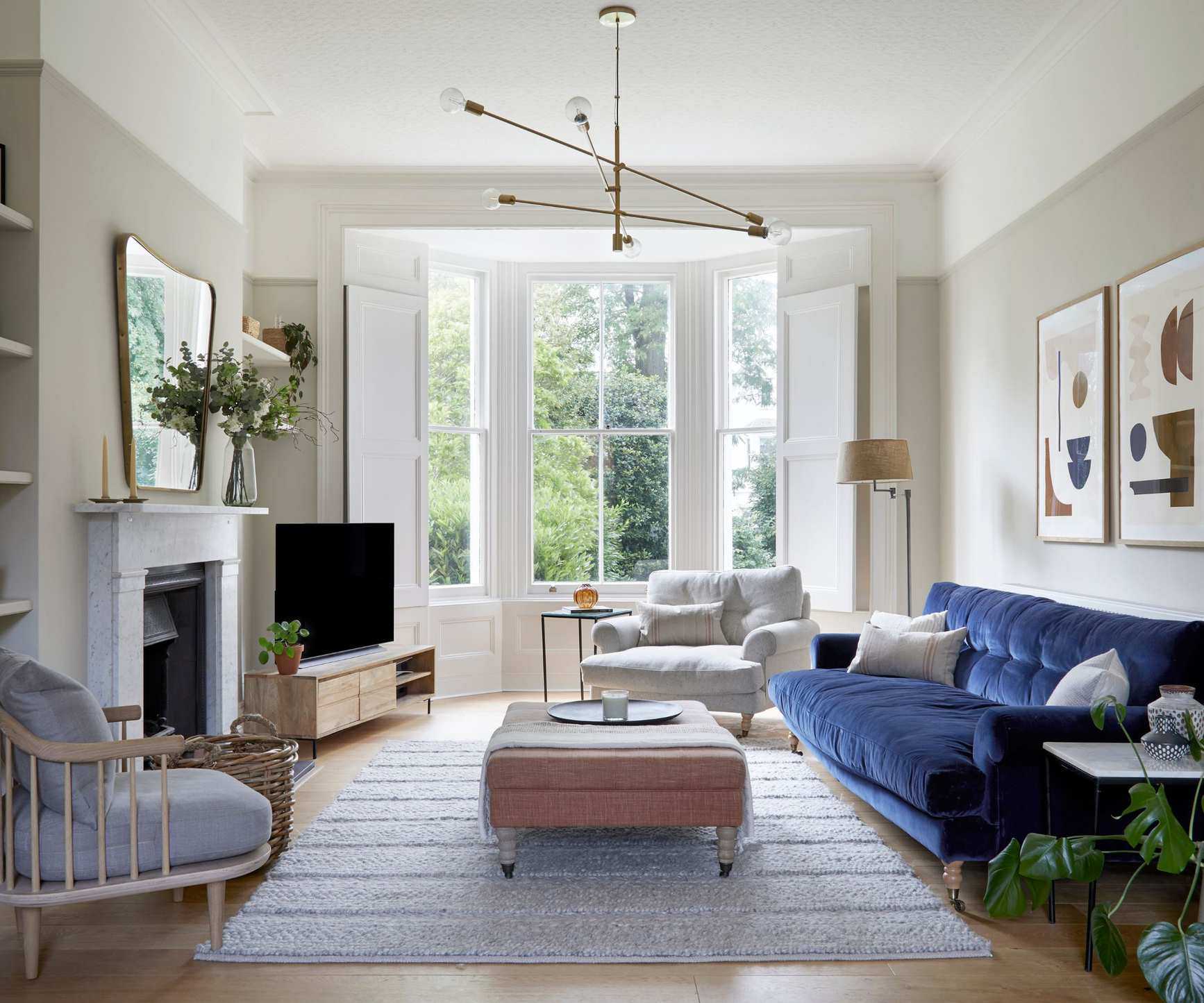 Living room with taupe walls, large bay window, grey rug at the centre, a pink ottoman on top, dark blue velvet sofa, and a large fireplace with a mirror above it