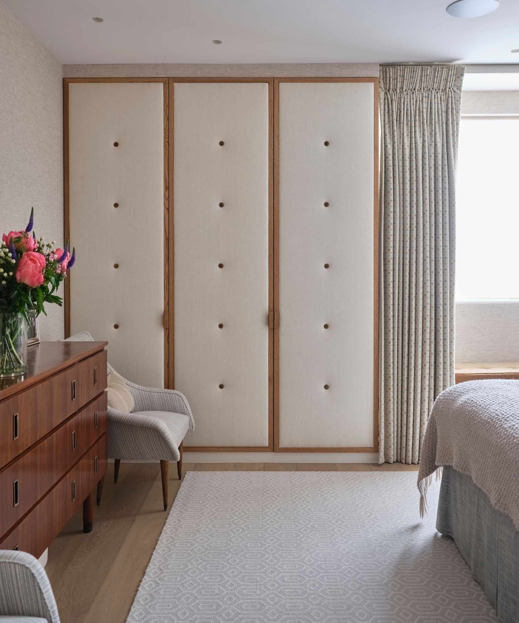 A cream and wood padded closet in a bedroom with a cream rug, wooden sideboard with vase of bright pink flowers, and grey chair.