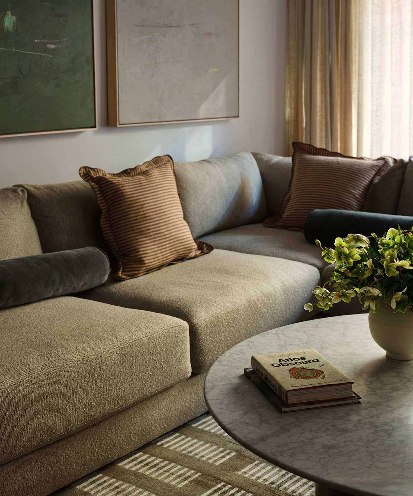 Living room with brown boucle sectional sofa, green geometric rug, a round marble coffee table with books and a plant on top, and two large paintings above the sofa in green and beige