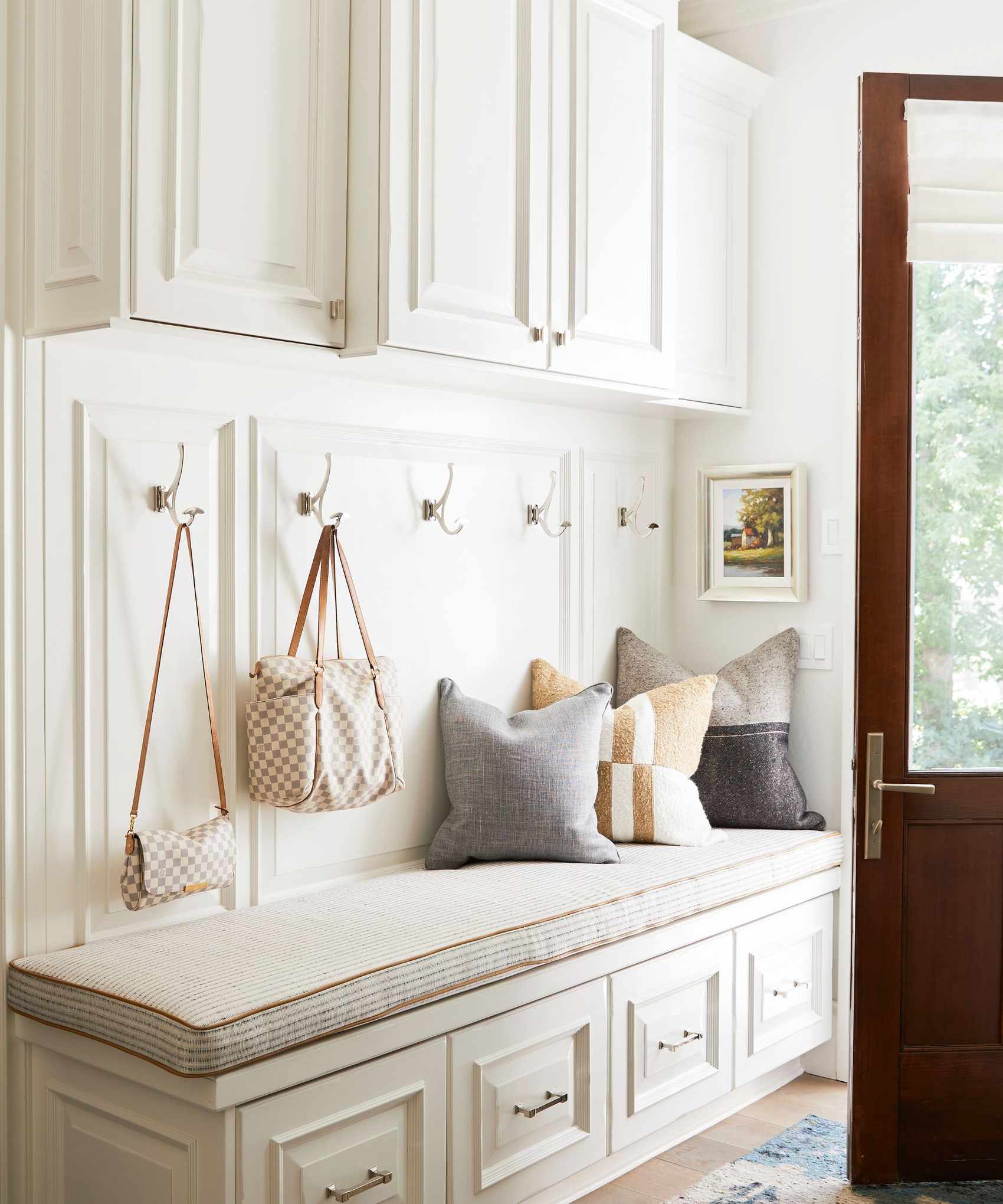 A white built-in storage bench in entryway with a padded top and cushions. Bags hanging from hooks above.