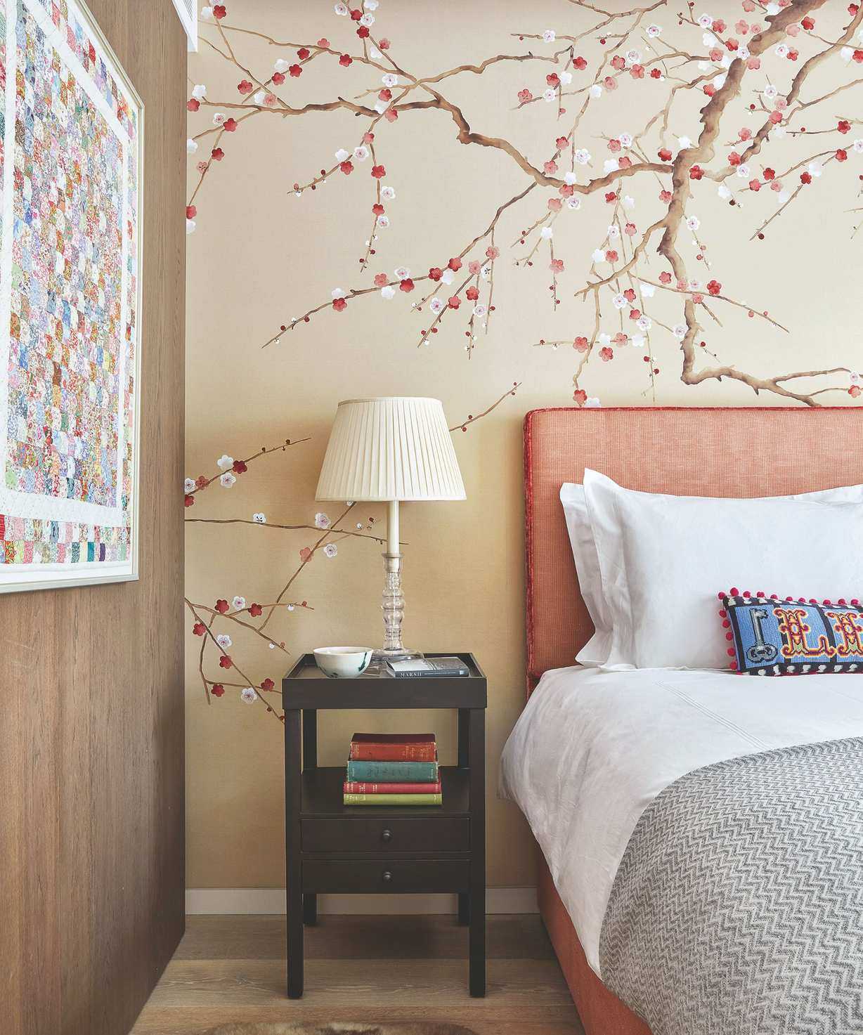 A bedroom with an orange bed to the right with white bedding, a grey blanket, and colorful cushion. The wall behind the bed is a neutral beige color with a cherry blossom tree motif. To the left of the bed is a black bedside table with a stack of books and white lamp, and to the left of that is a wood panelled wall with a bright and colorful abstract artwork. The floor is wooden.