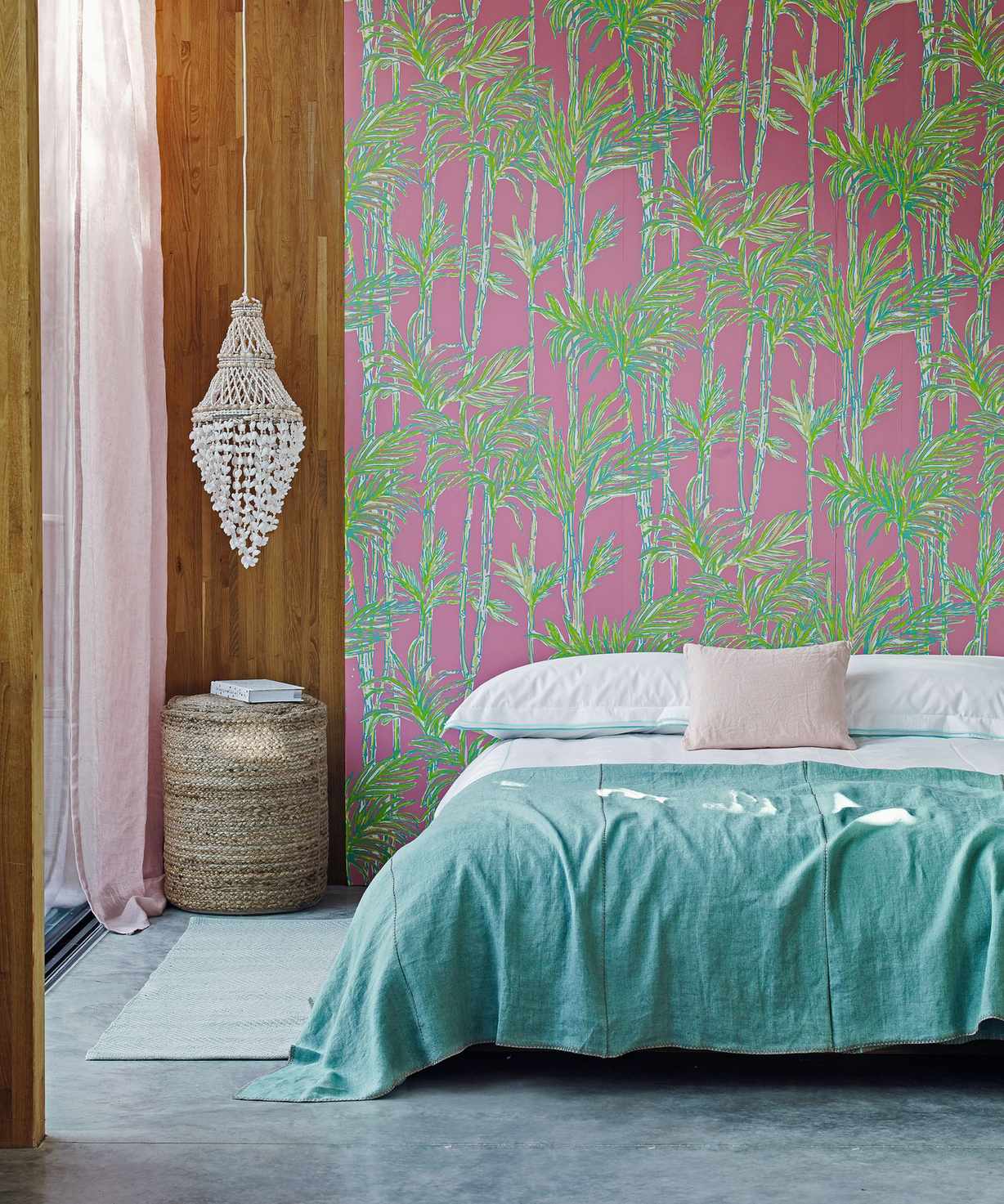 A colorful bedroom with a pink and green patterned statement wall, wood panelling, a macrame hanging, rattan stool, and a low bed with blue bedding.