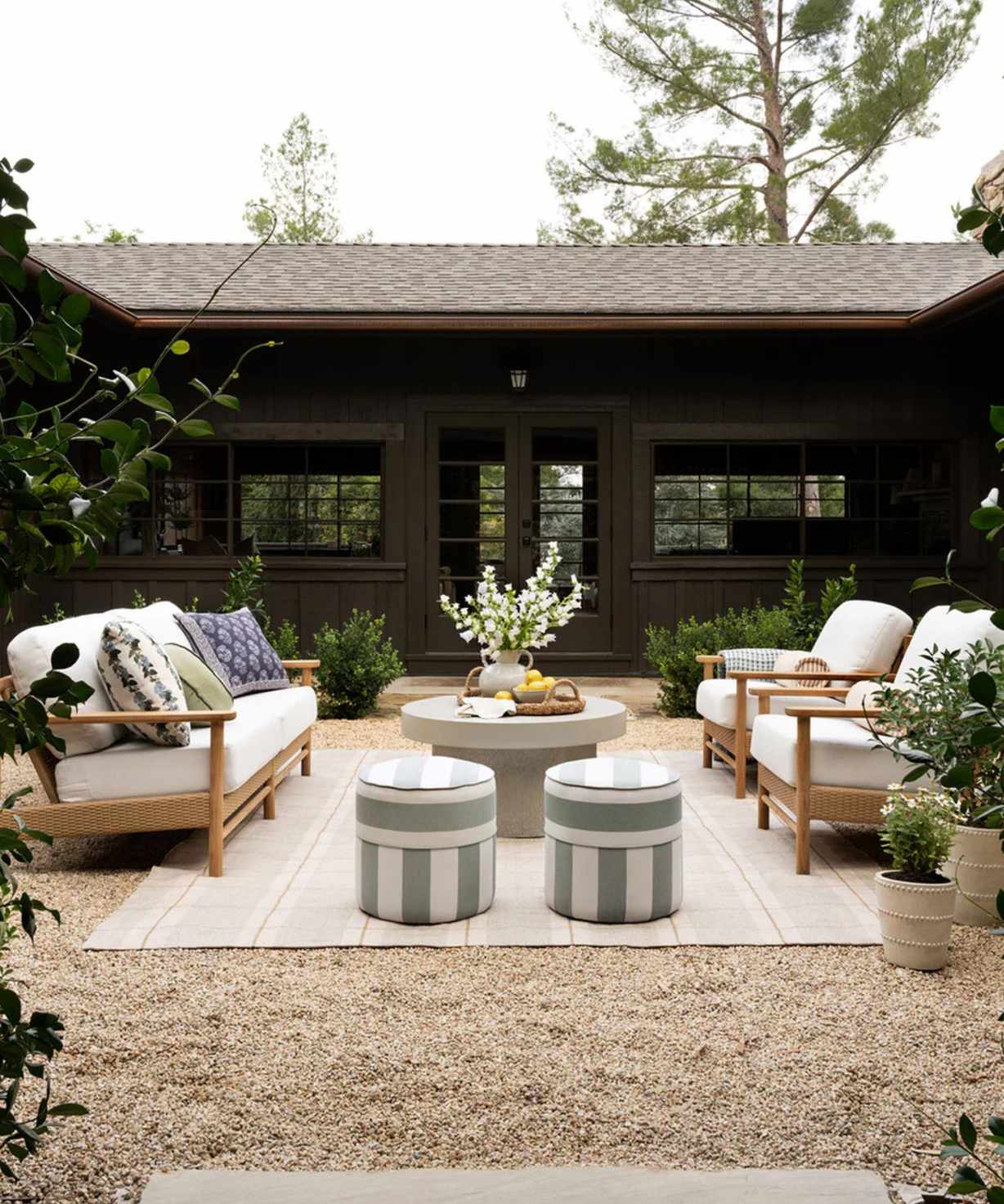 mcgee &amp; co outdoor living room in a shingled courtyard with wooden sofas facing each other on a striped rug with two small green and white striped ottomans
