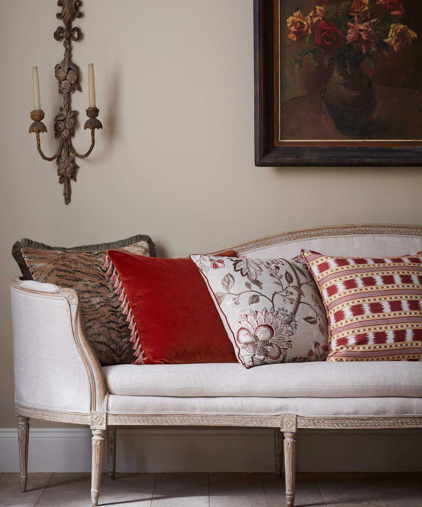Vintage looking white upholstered chaise with four throw pillows in different patterns and deisgns in white, red, brown and taupe. There is an ornate wall-mounted candle holder with slim candles, and framed art on the wall with autumnal blooms
