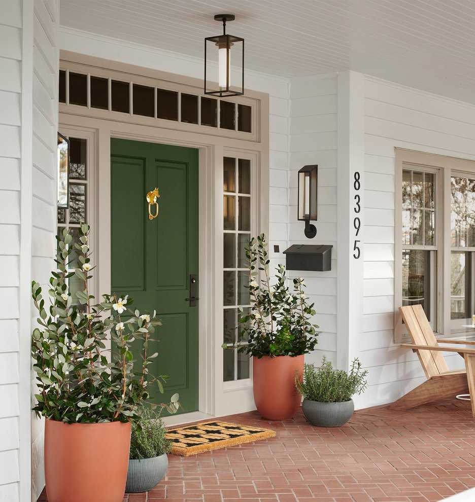 Traditional home front porch with green door, potted plants and a welcome mat. There is an Adirondack chair in view to the right and and the floor is red brick tile.