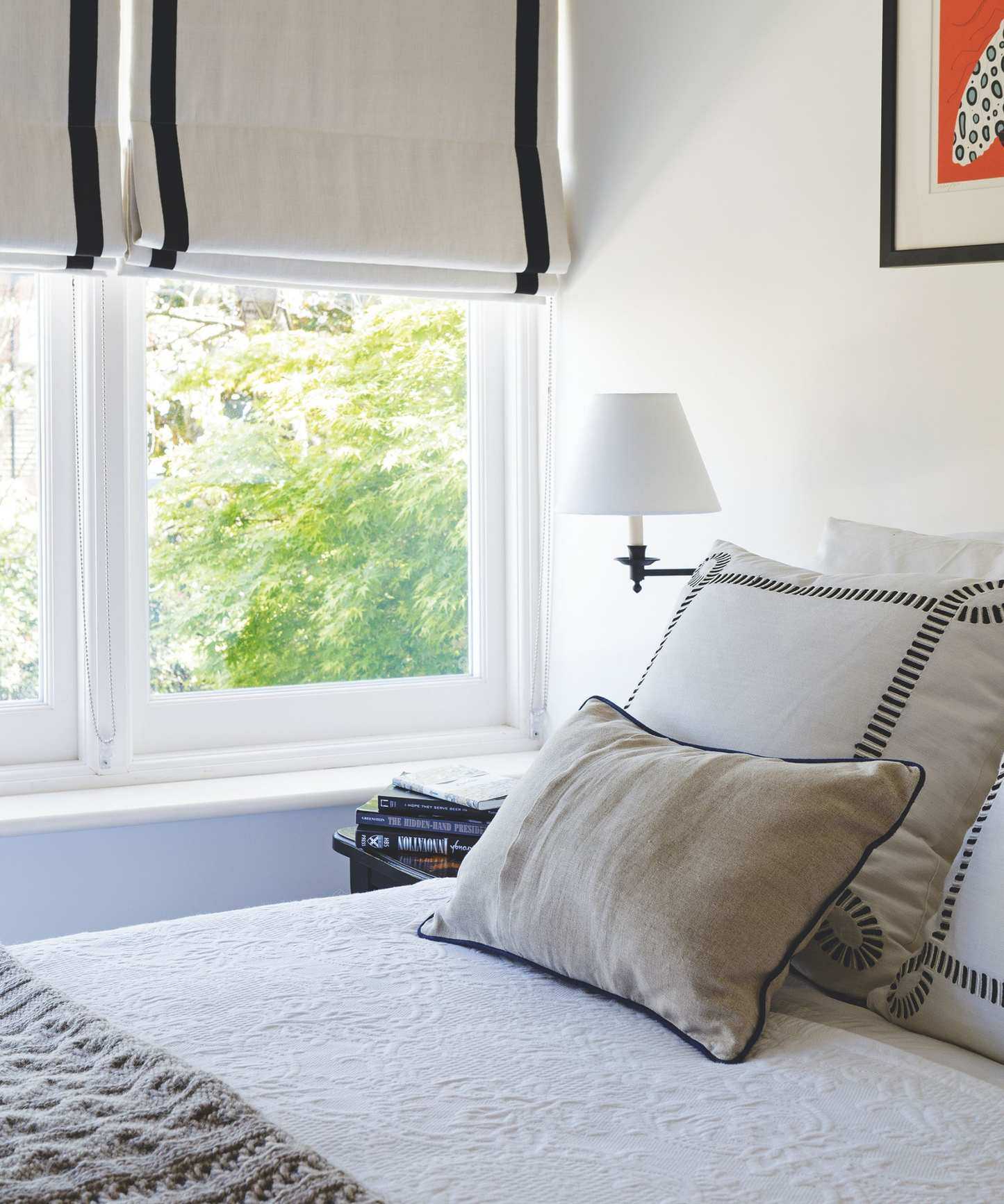 A white bedroom with a bed with white bedding in the foreground, with a grey cushion at the foot and two large square white and black cushions at the head, with a rectangular neutral beige cushion leaning up against them. To the left of the bed is a white wall lamp, and the corner of a bedside table with a stack of books on it is seen. Behind is a large window with green leaves seen beyond, and a cream and black blind.