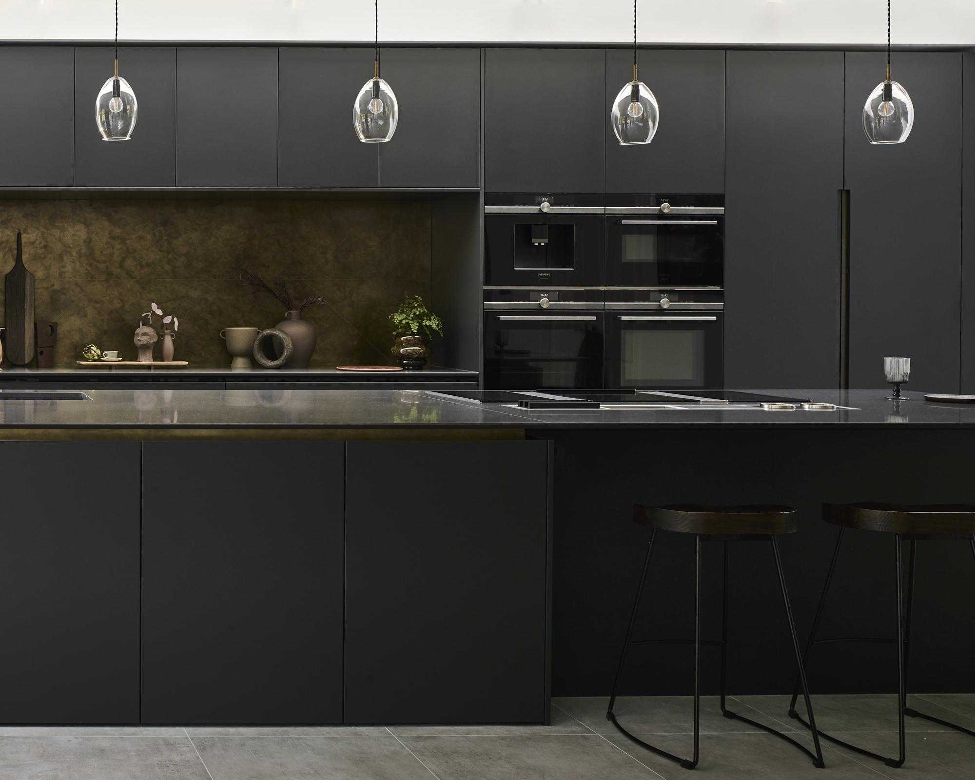 A black kitchen idea with matte black cabinets, stone worktop on the island, brass backsplash and pendant lights