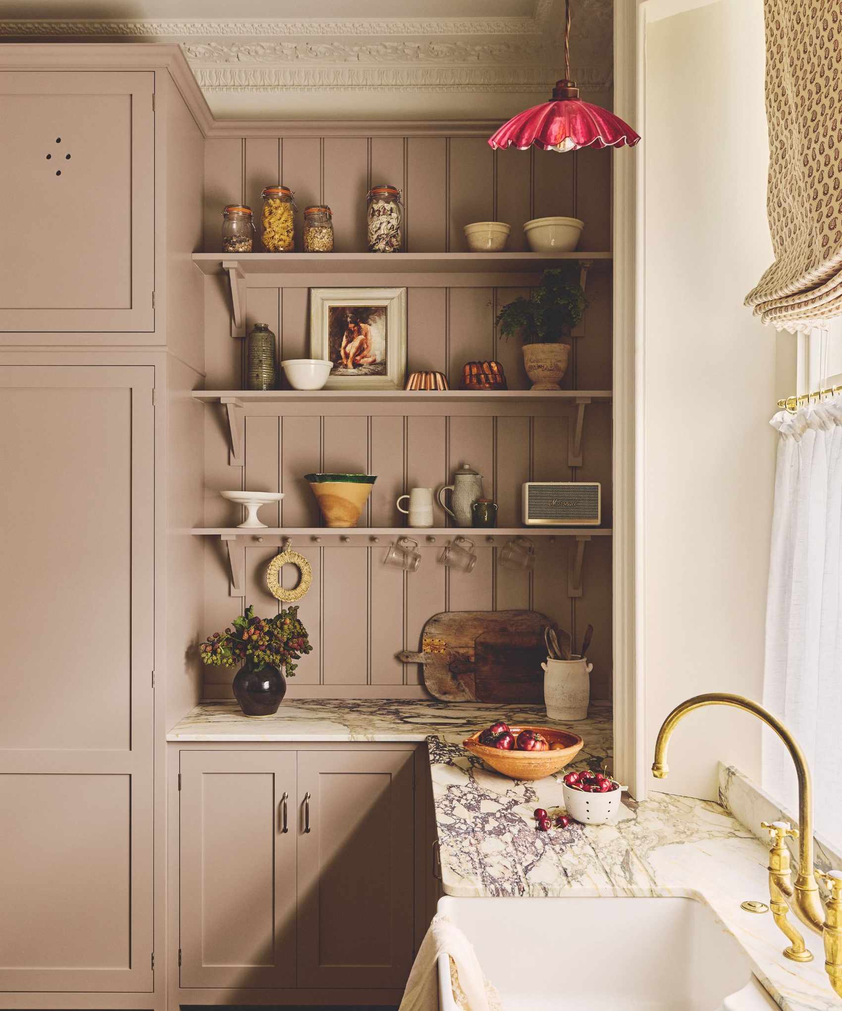 dusty pink lilac traditional shaker kitchen with marble countertops, open shelving with vintage decor, a cafe curtain window and roman blind and a sink with brass faucet