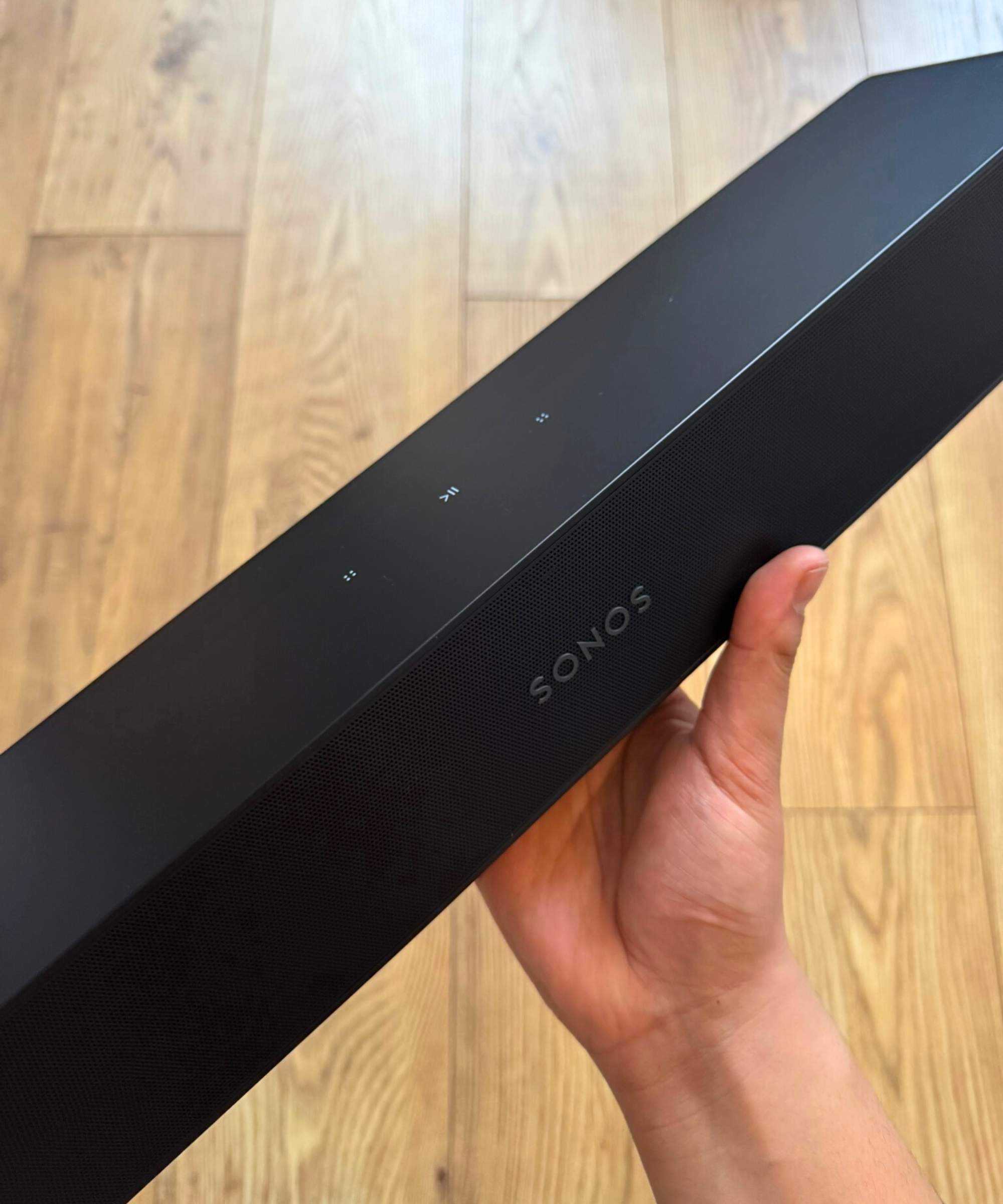 Hand holding the Sonos Ray soundbar with wooden floor in background.
