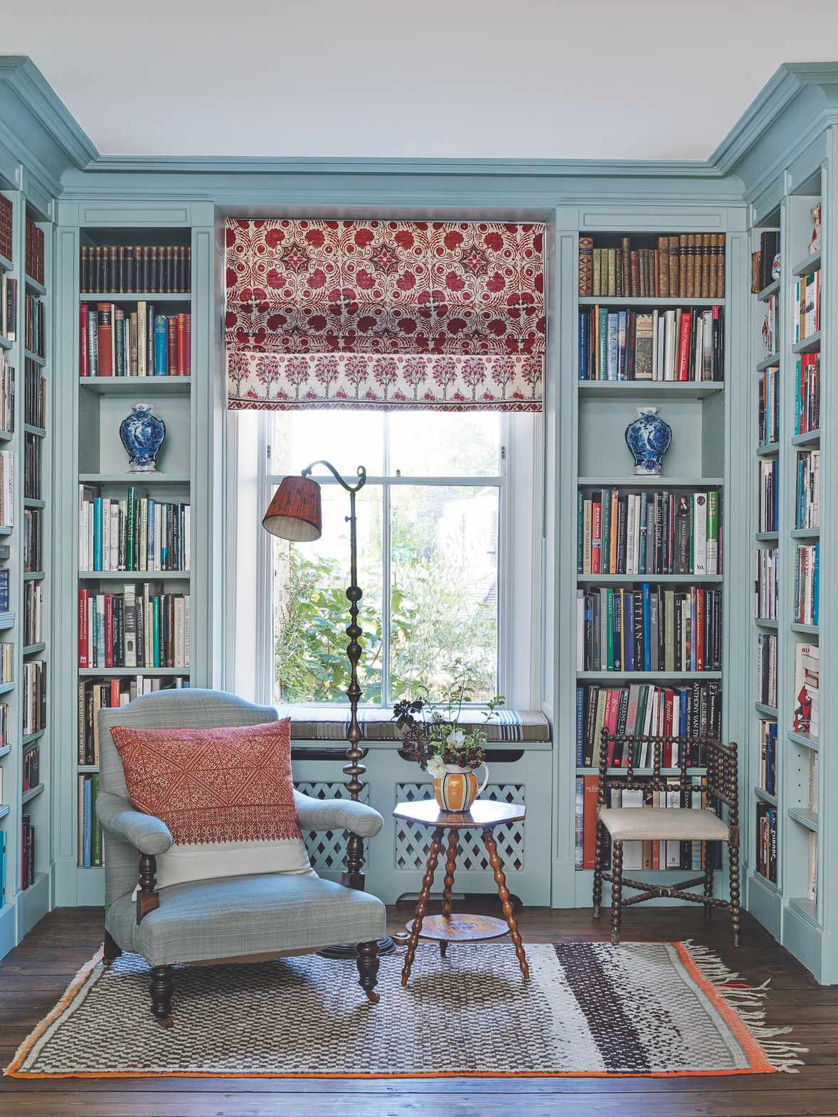 Home library with blue bookcases around the walls, wood floor with rug, blue armchair and window with red patterned Roman blind