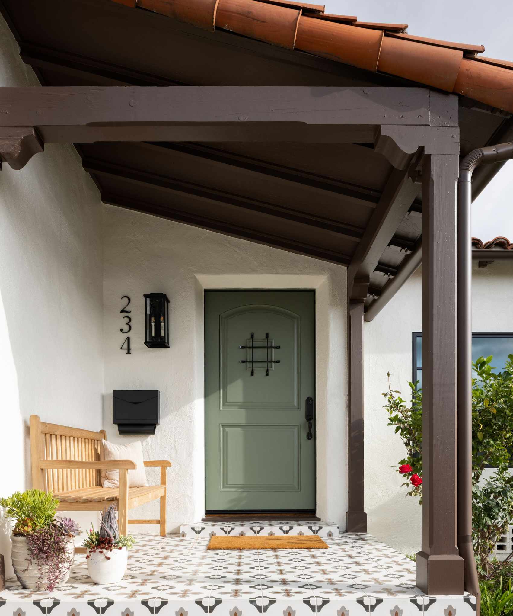 Spanish style home with terracotta tiled roof, white and black tiled floor, green door, wood bench and welcome mat