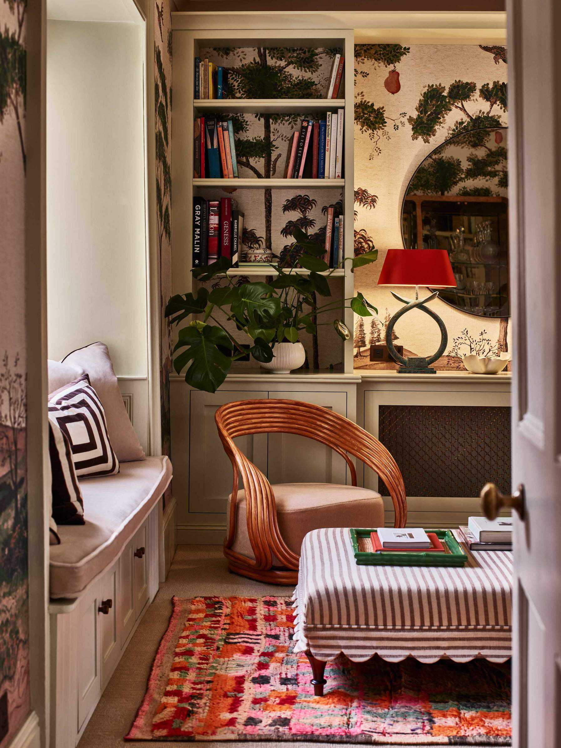 cozy snug room with mural wallpaper and neutral bookshelves styled with a red table lamp, window seat nook with a striped ottoman with scalloped trim