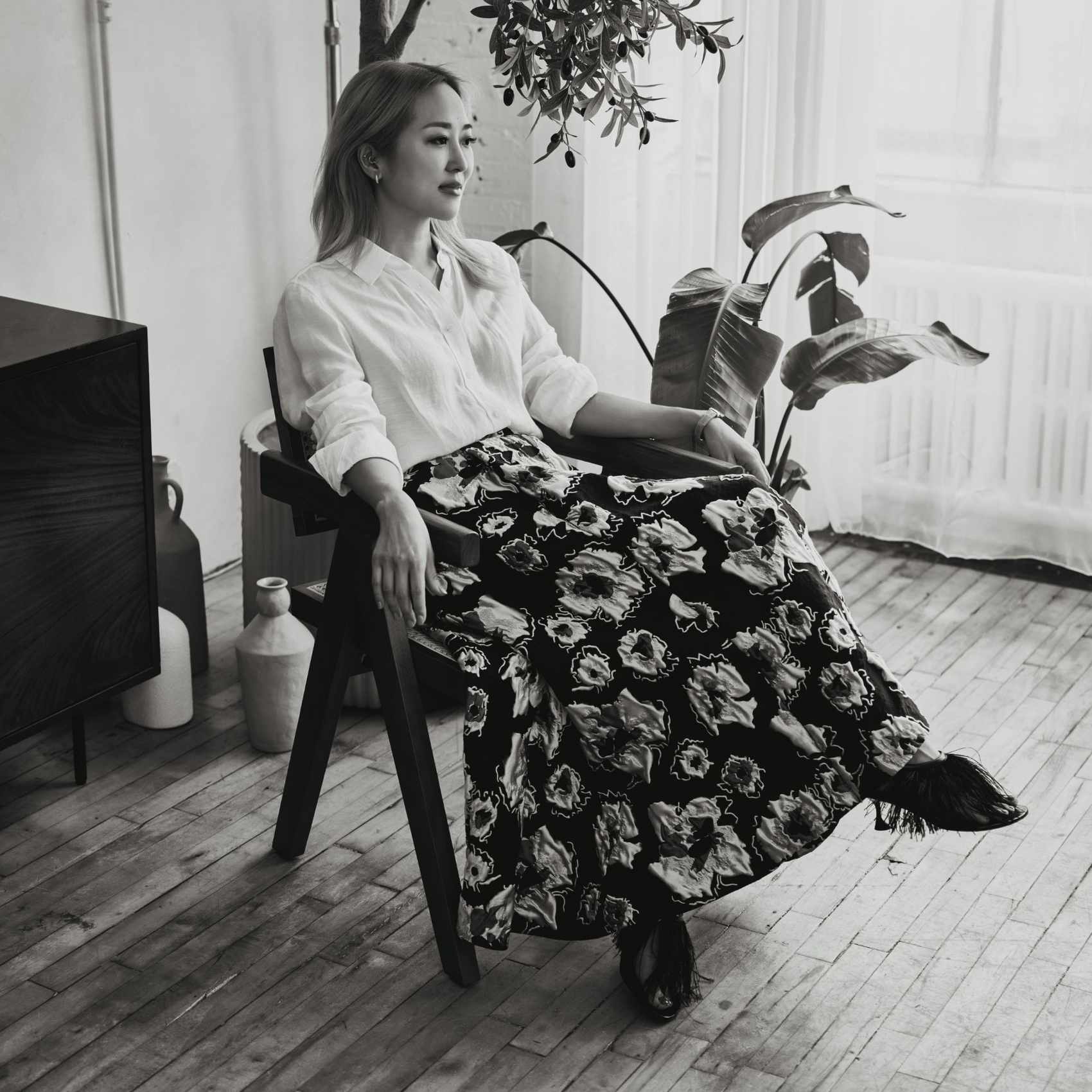 A black and white image of Maiko Shimazaki wearing a white skirt and black and white patterned skirt, sitting in a chair.