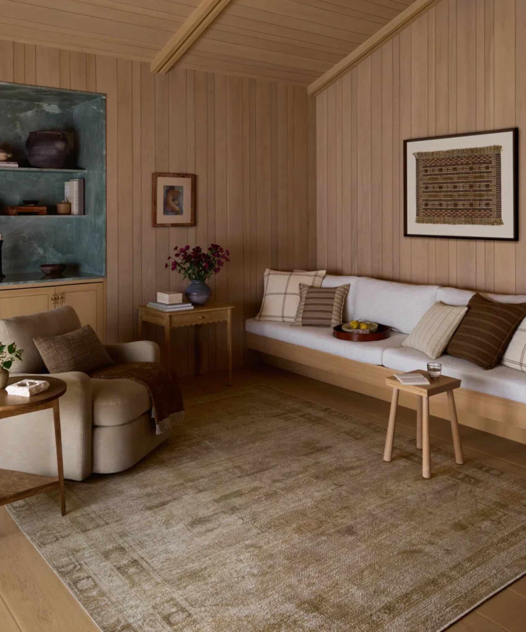 a cozy living room with a large area rug, a built in couch, wood-paneled walls, and a taupe accent chair