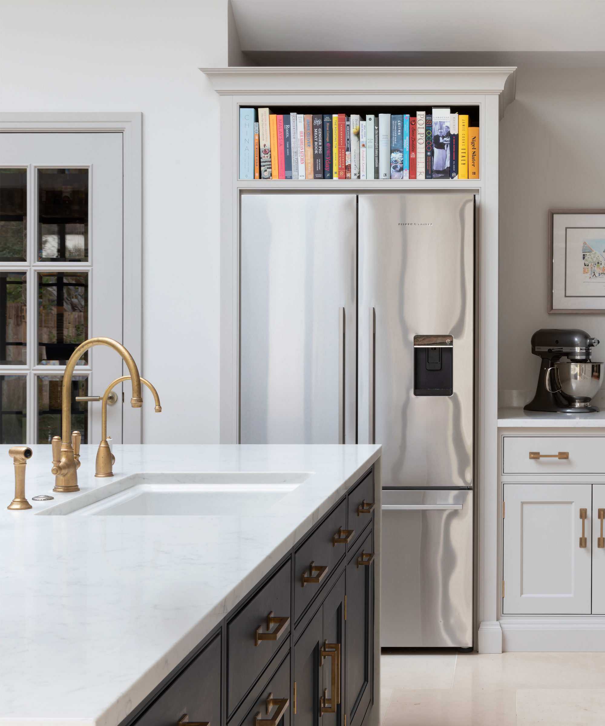 Close up of corner of kitchen with large double fridge in alcove, shelving and cabinetry surrounding, kitchen island with sink, brass tap