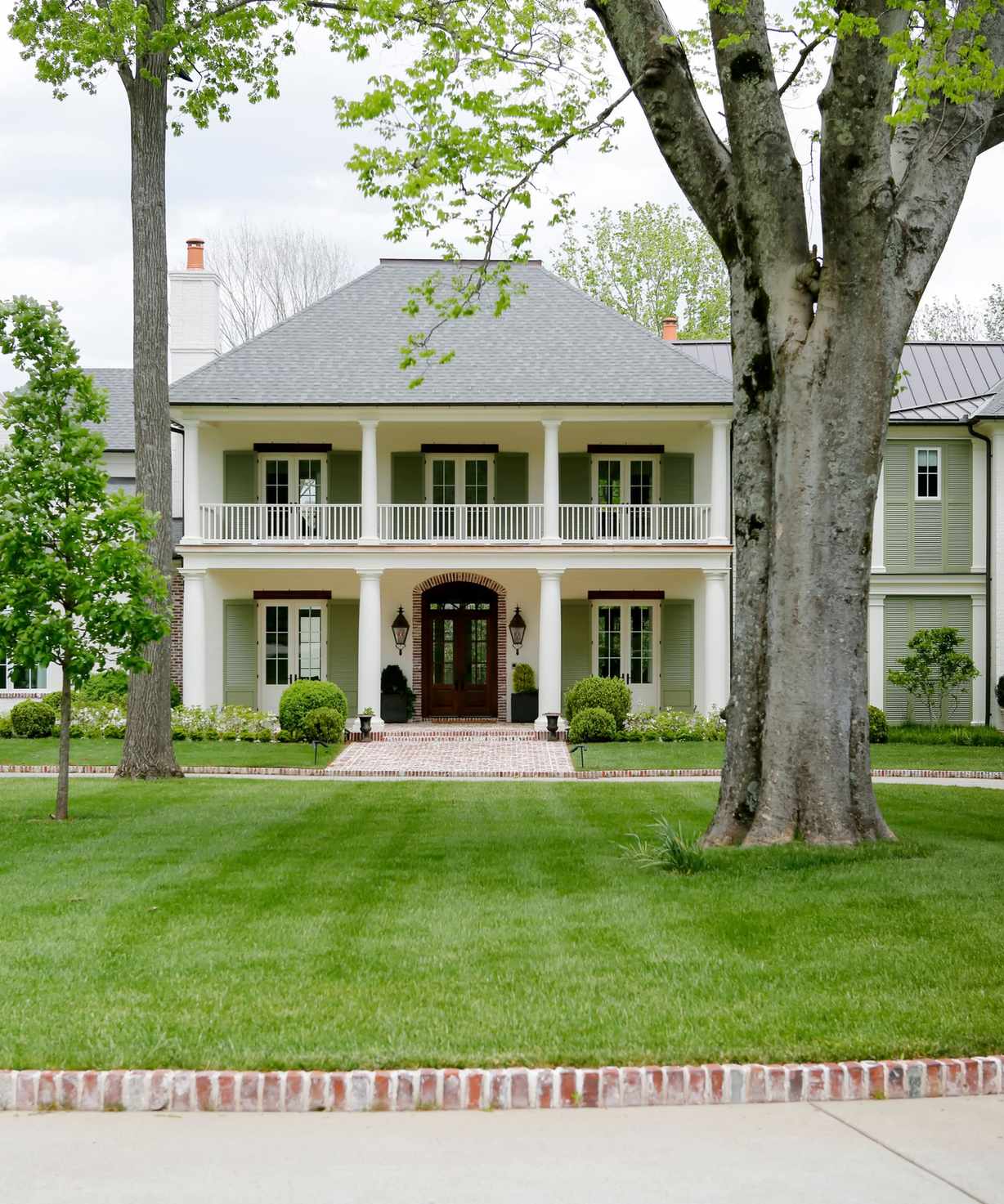 A traditional-style house exterior with muted green and soft white paintwork, a lush green lawn with trees, and a paved path.