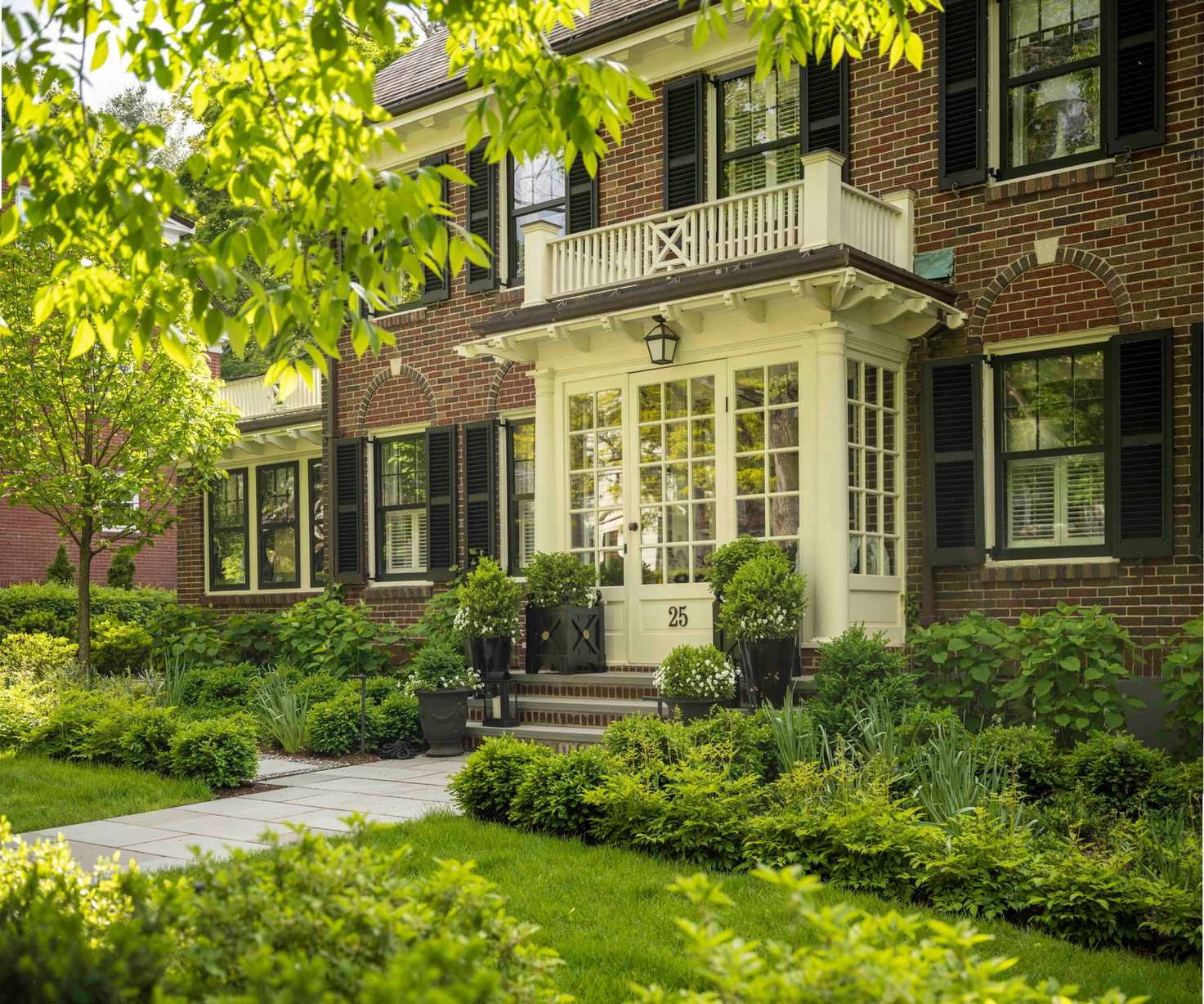 Grand red brick home with white balcony and front door and luscious greenery and potted plants