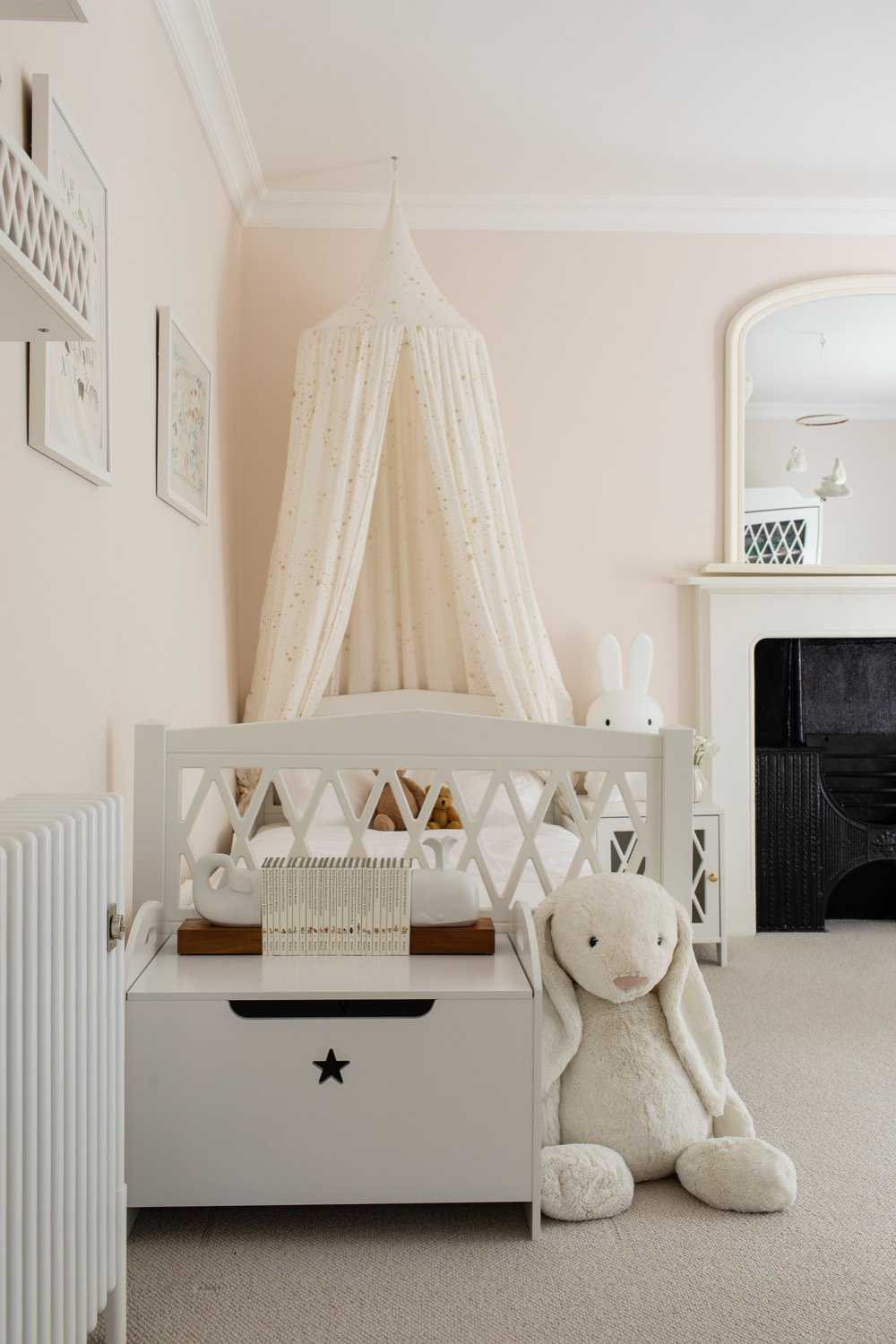 kids bedroom with very pale pink walls, fabric canopy over the bed, white painted bed, toy chest, fire place, mirror on mantel, artwork, toy