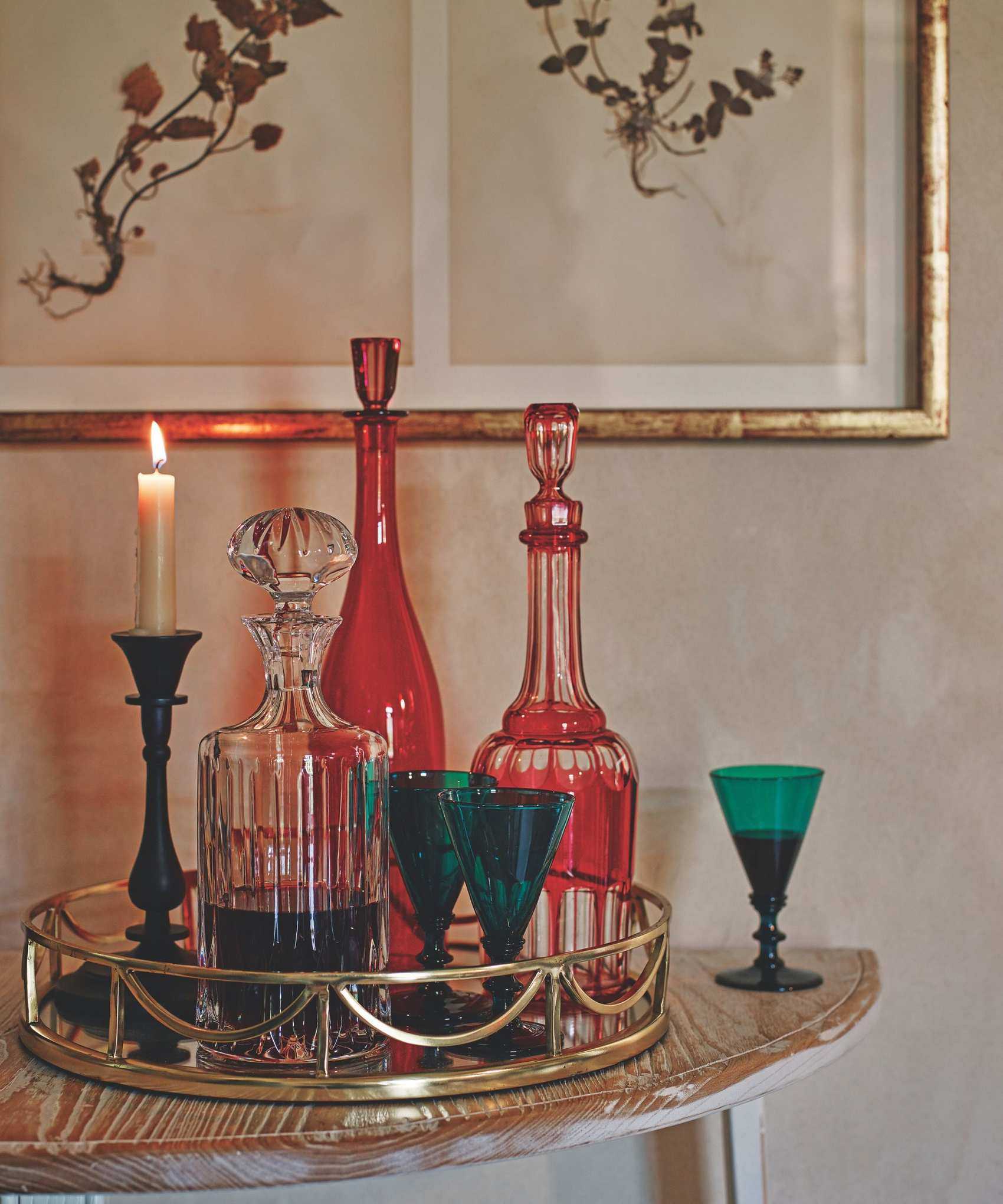 Vintage colored glassware on a gold mirrored tray on a wooden table. There is a lit taper candle, and above are two vintage-look botanical prints in a gold frame.