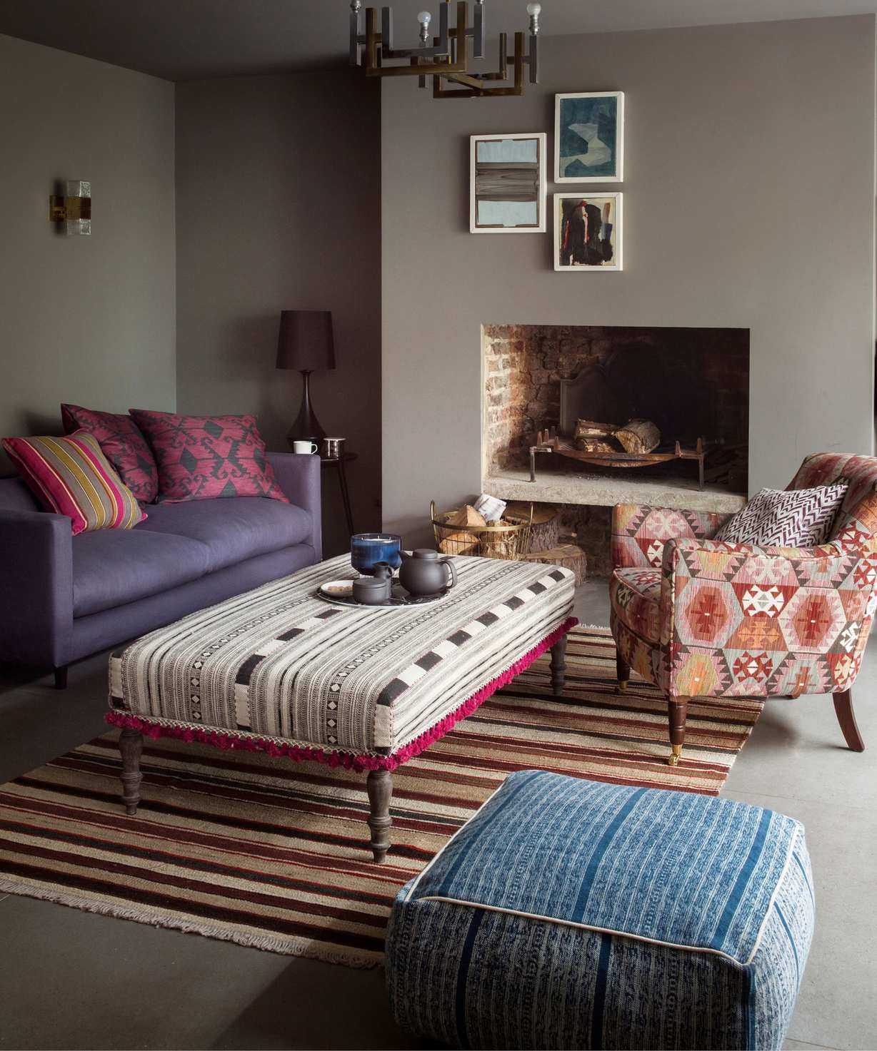 A neutral brown living room with contrasting patterned ottoman, chair and pouffe, striped brown rug and purple sofa with bright pink cushions and three framed pictures in white frames below a metal industrial pendant light