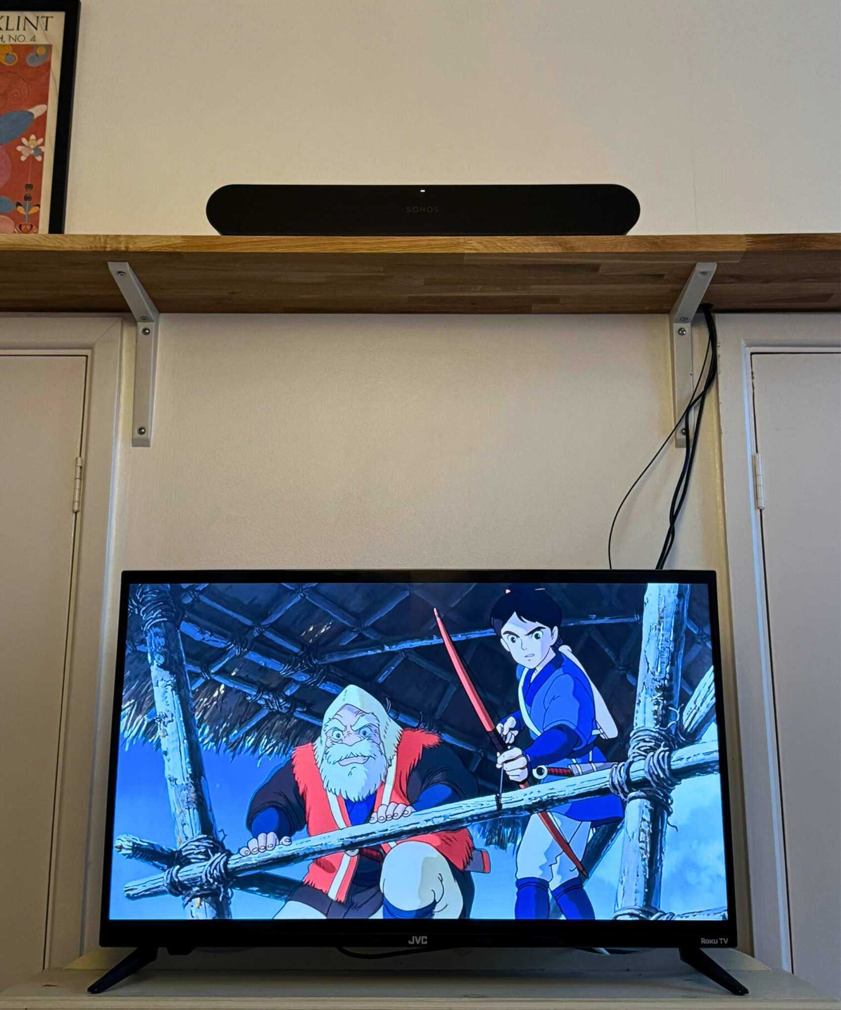 The Sonos Ray soundbar on a wooden shelf above a TV showing Princess Mononoke, in front of cream-white wall.