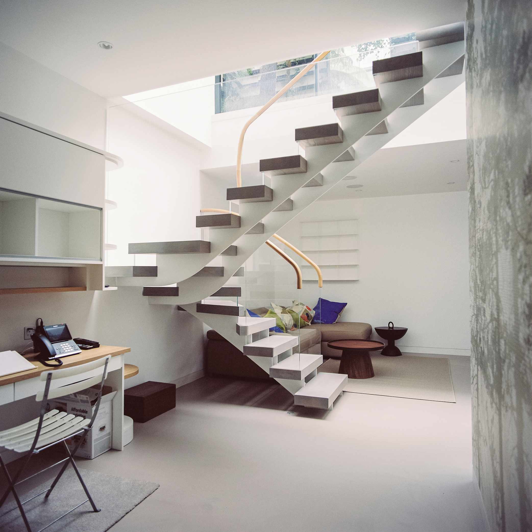 Open tread staircase with glass balustrade leading to basement with neutral decor with seating area and desk