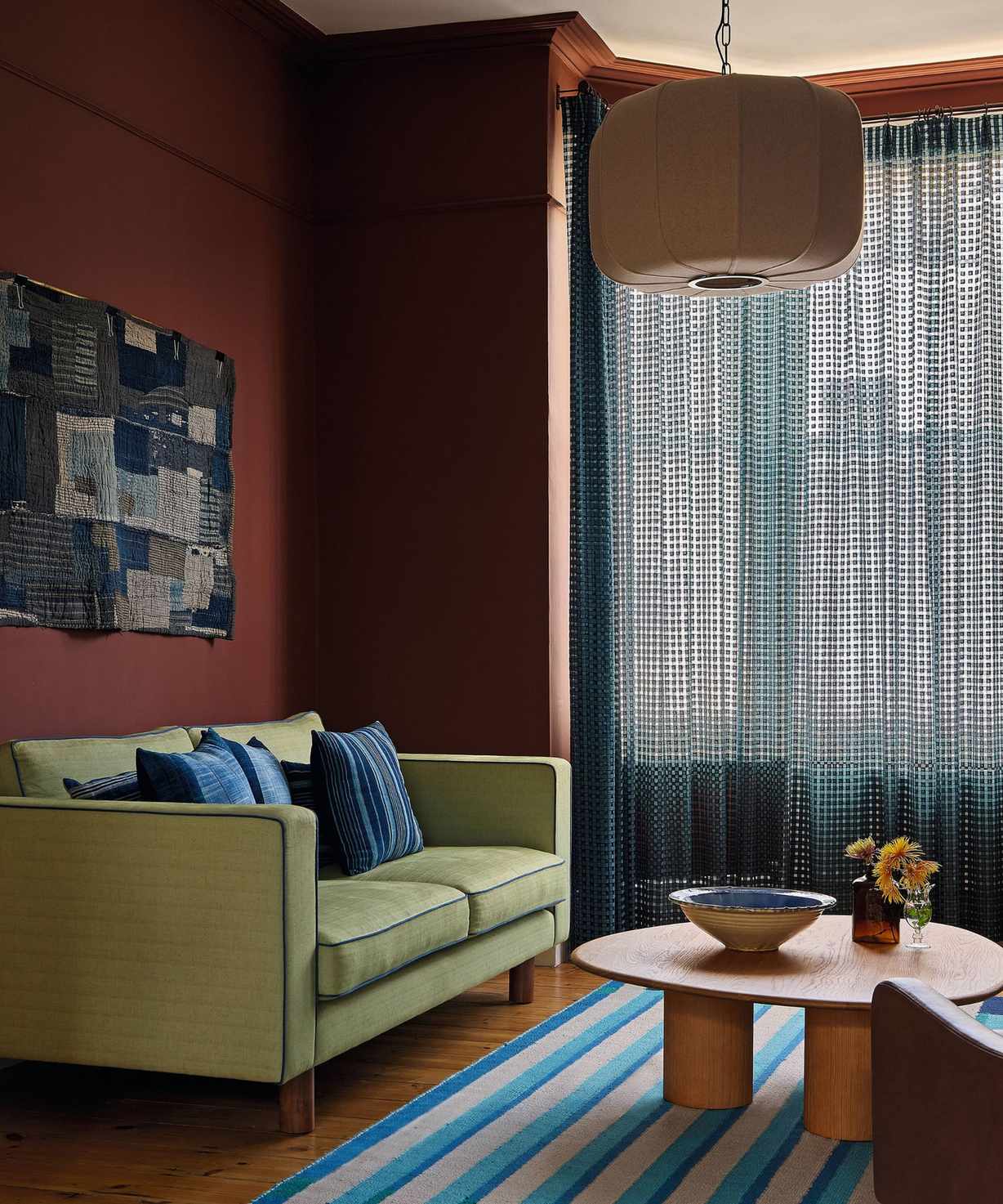 a red painted victorian living room with high ceilings and a bay window. a lime green couch is styled with a blue striped rug and blue sheer drapes at the window