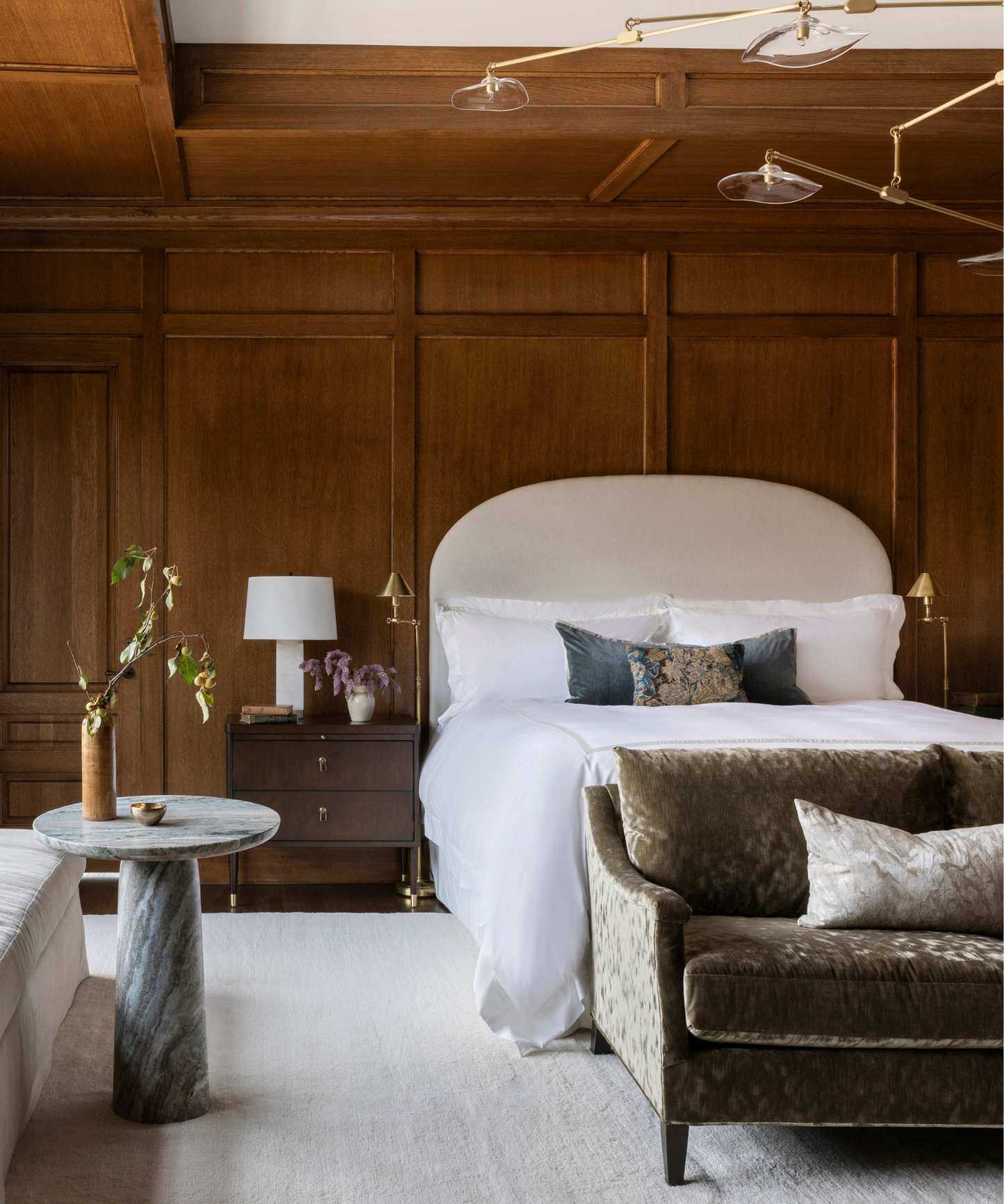 calm timeless bedroom with original dark wood panelling, a large neutral rug, neutral bed, dark green velvet couch at the foot of the bed and a small marble table