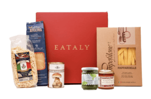 A terracotta colored box is pictured at center. On either side are components of pasta dinners, including three bags of pasta.