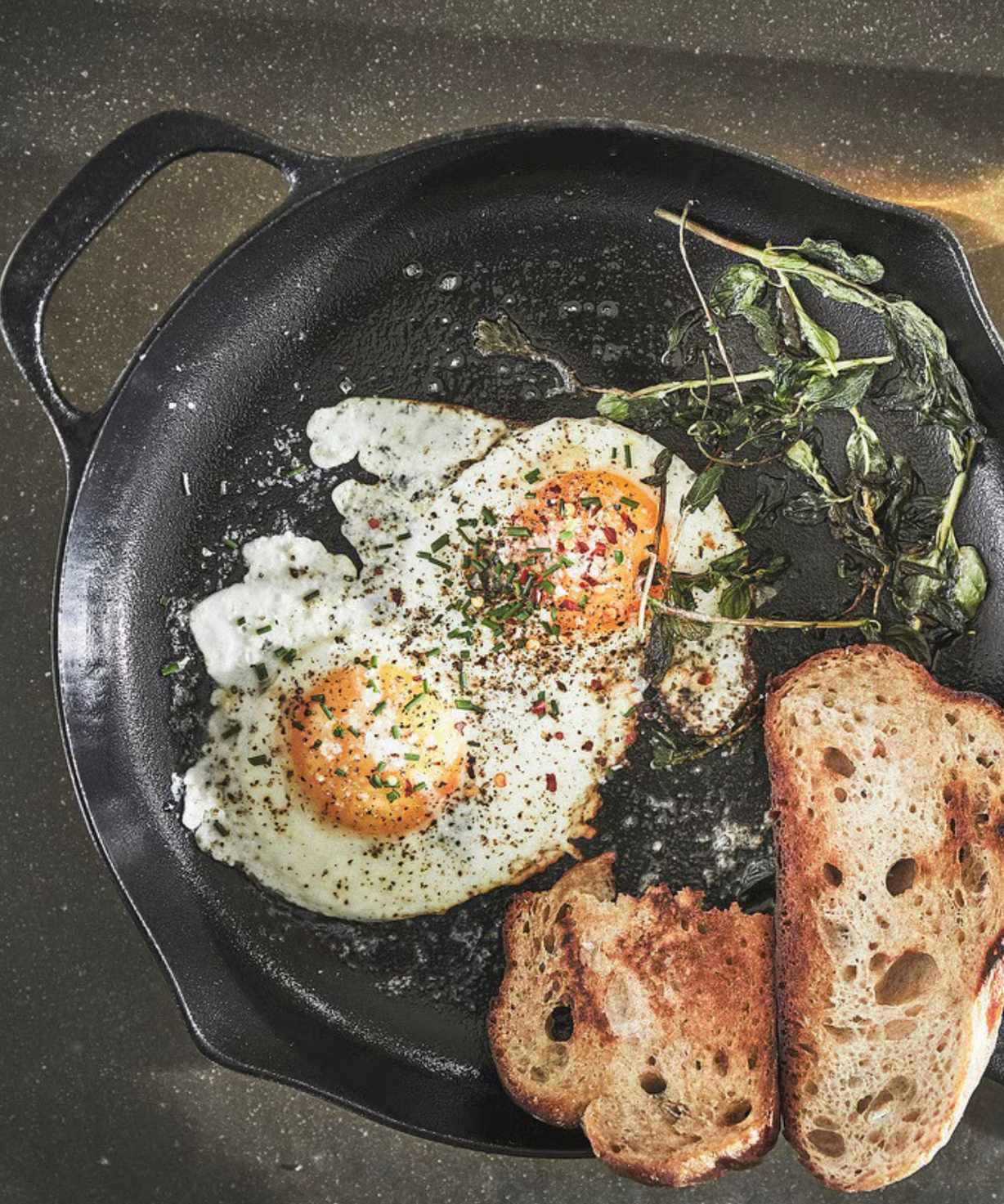 A black cast iron skillet on a black worktop, with two fried eggs, two slices of rustic toast, and some wilted spinach inside.