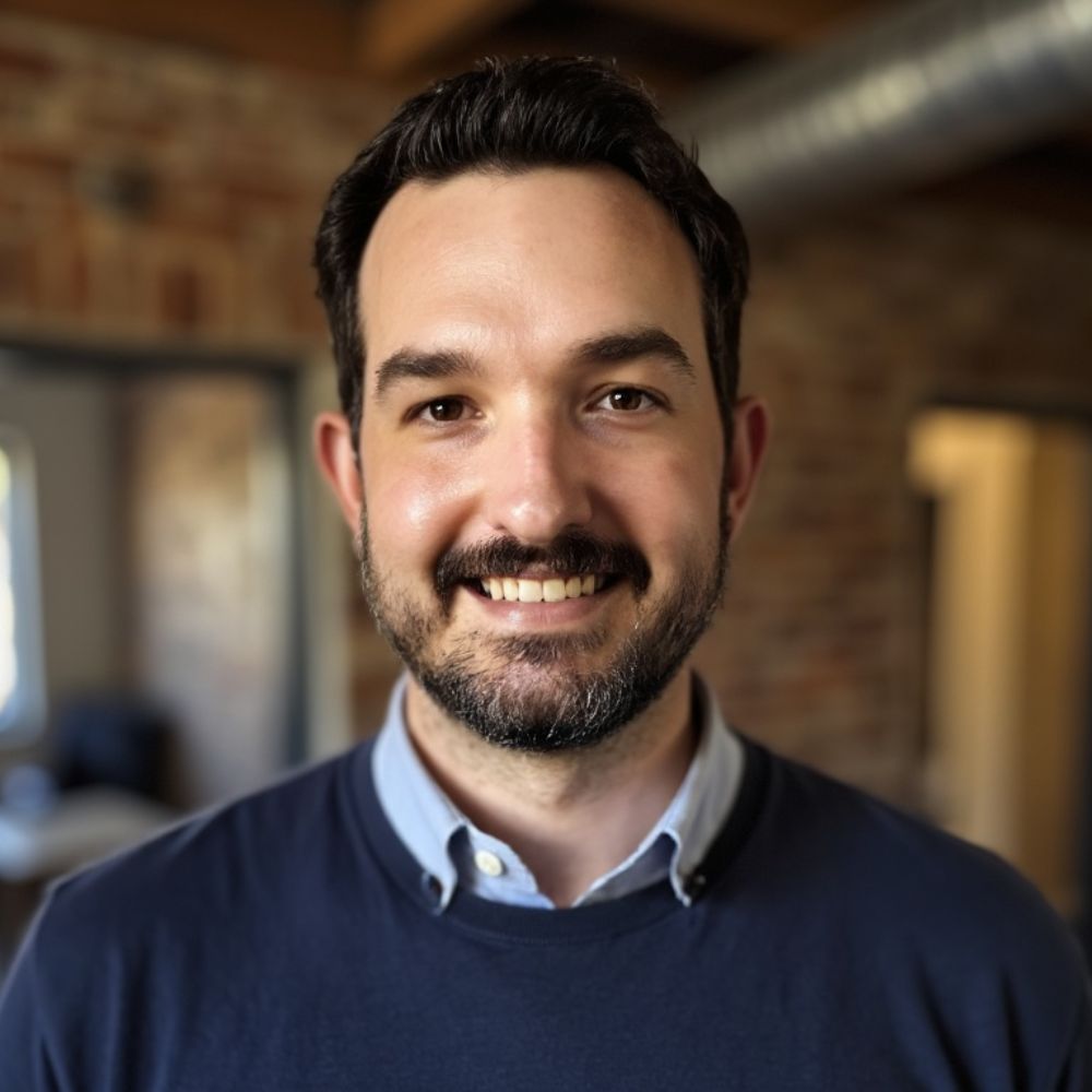 A headshot of Jon Riding wearing a blue shirt and blue jumper
