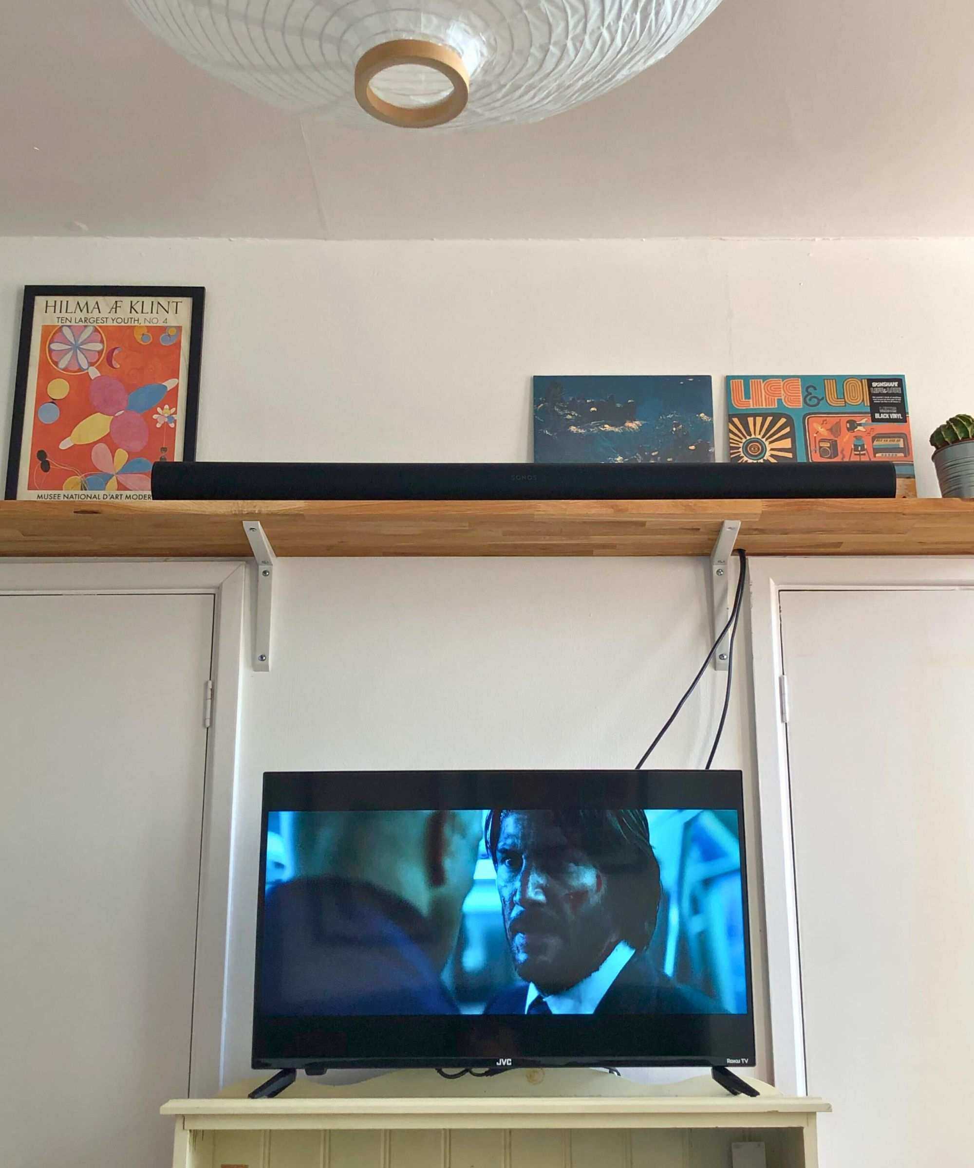 Sonos Arc Ultra soundbar in Dan Fauzi's home, on wooden shelf with cactus plant, above TV showing John Wick: Chapter 2. A white lampshade is pictured in the top left corner, above two prints on the wall.