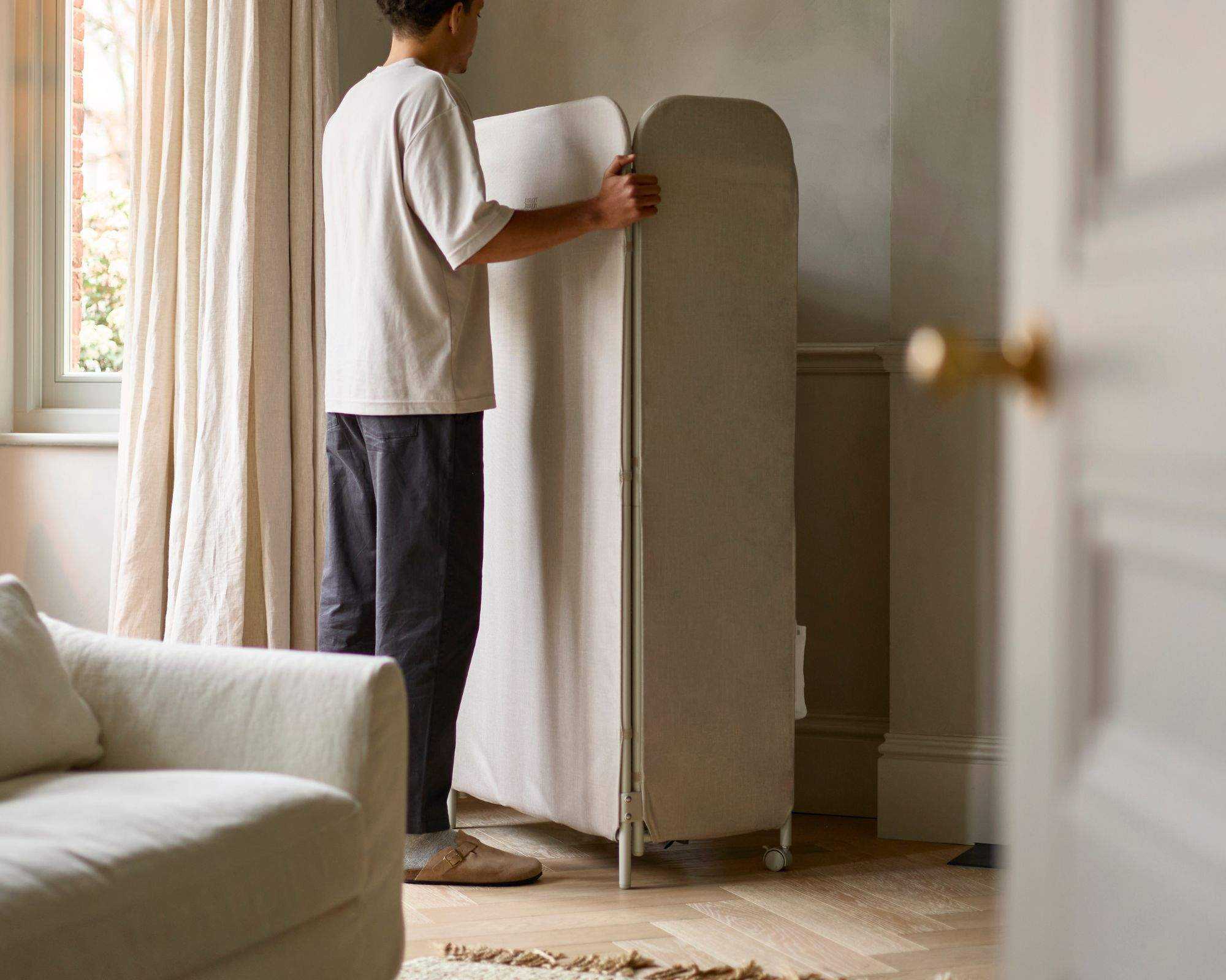 A man next to the Eclipse&trade; 3-tier Ecru Clothes Drying Rack With Screen in a bright living room
