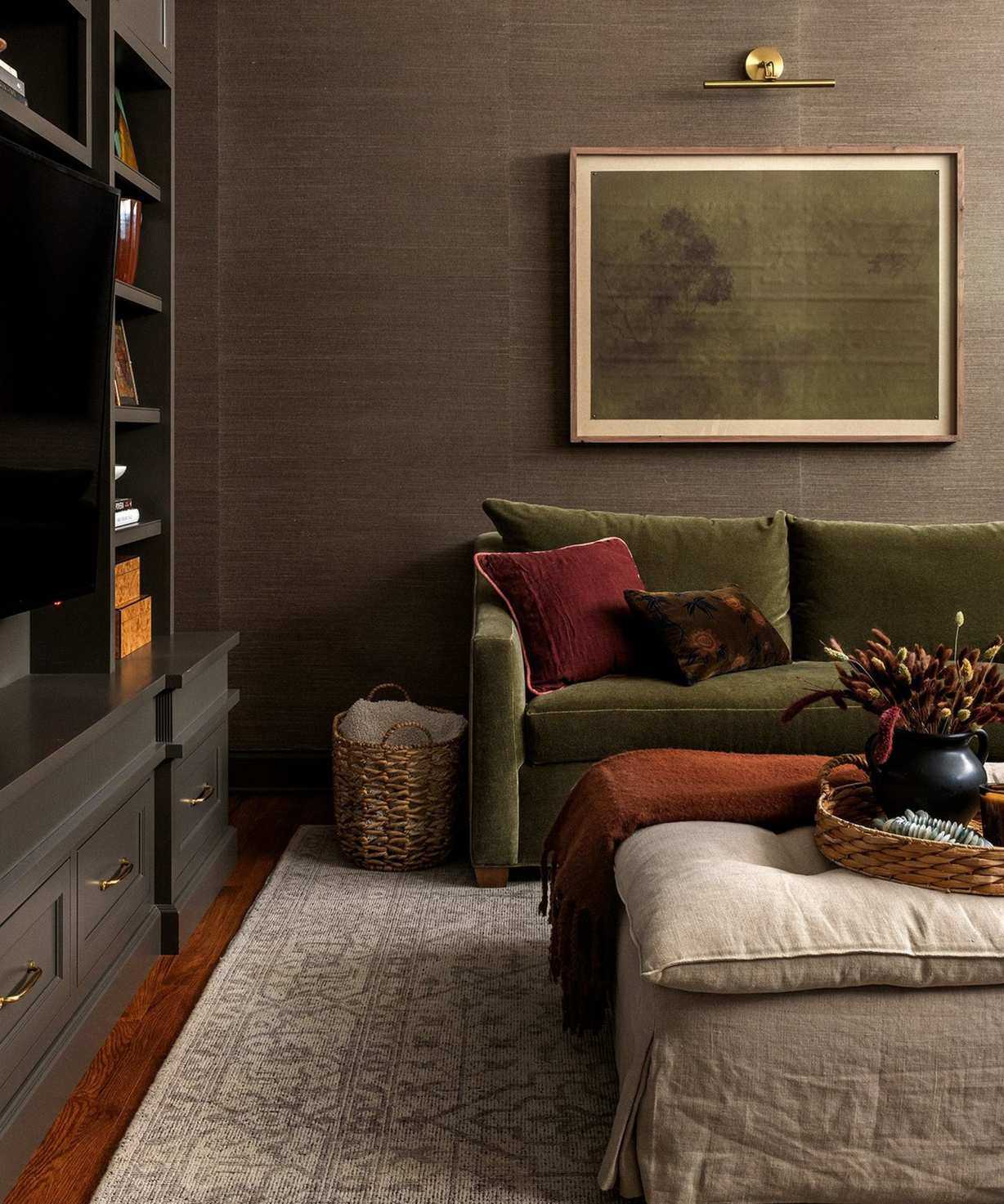 Dark living room with patterned rug, green sofa with cushions, framed minimalist painting, partial black bookshelf to the left, footstool, wooden floors.