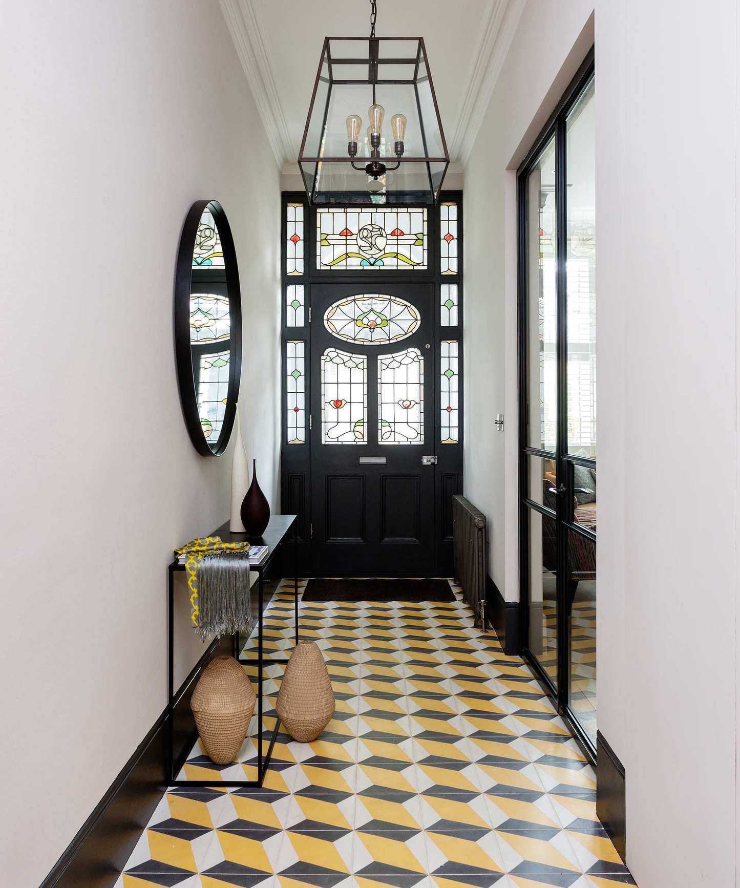 black and white entryway with black woodwork and door with stained glass, black round mirror and console, yellow and black 3d floor tiles, lantern light, crittal doors to living room