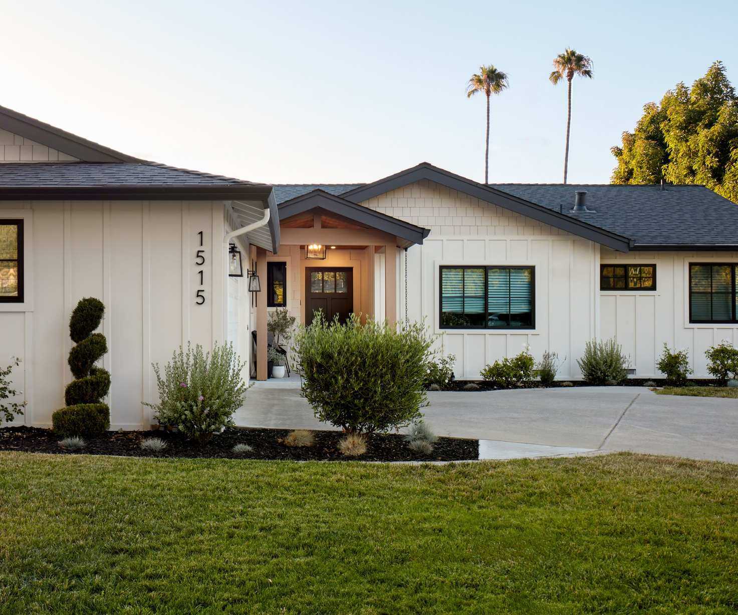 A bungalow home exterior with warm white siding, a dark brown door, and a light brown porch. Dark window trim and a gray roof with a grass lawn and paved driveway.