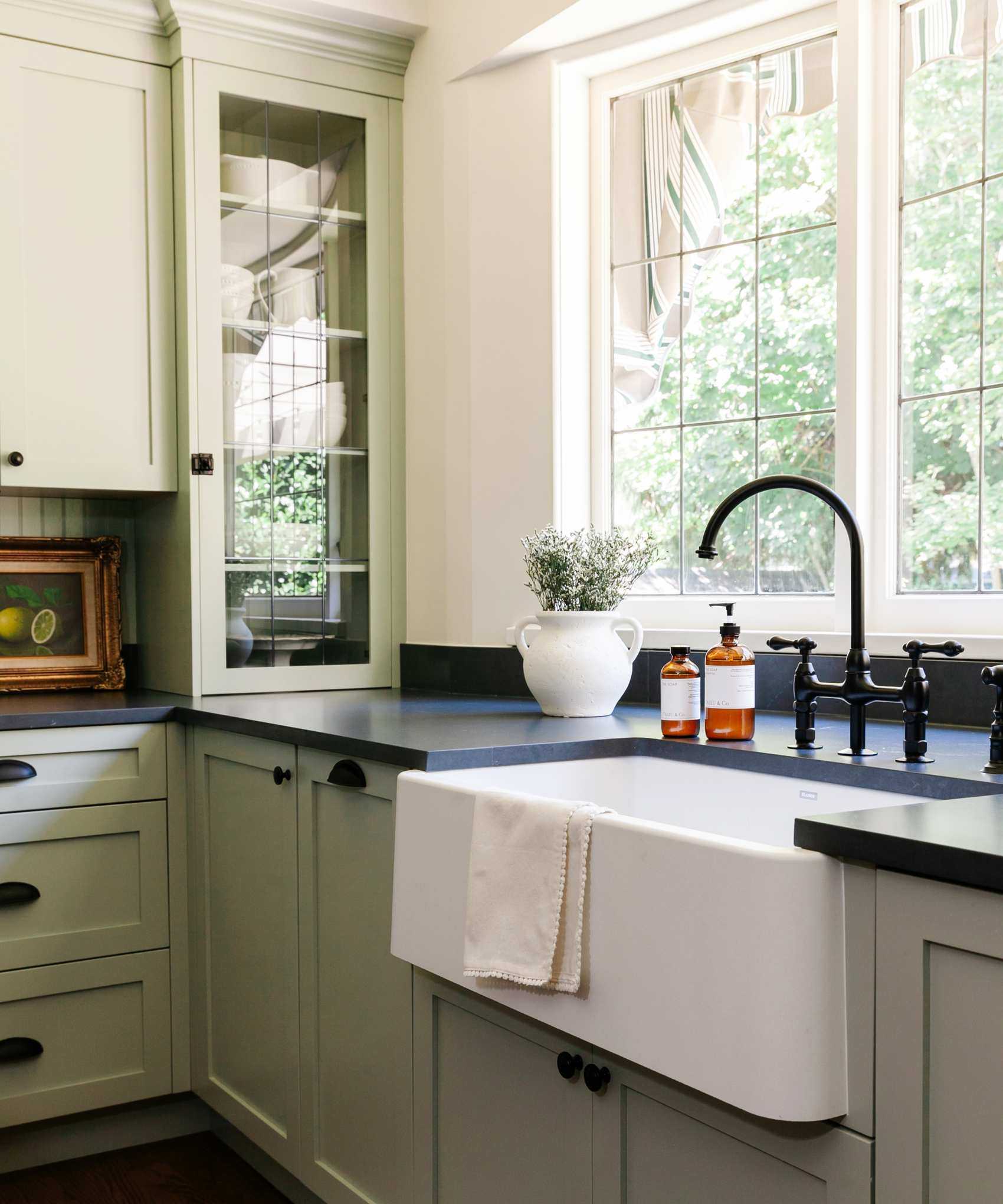 A sage green kitchen with large windows, a white basin sink, and a small plant sitting on a black countertop