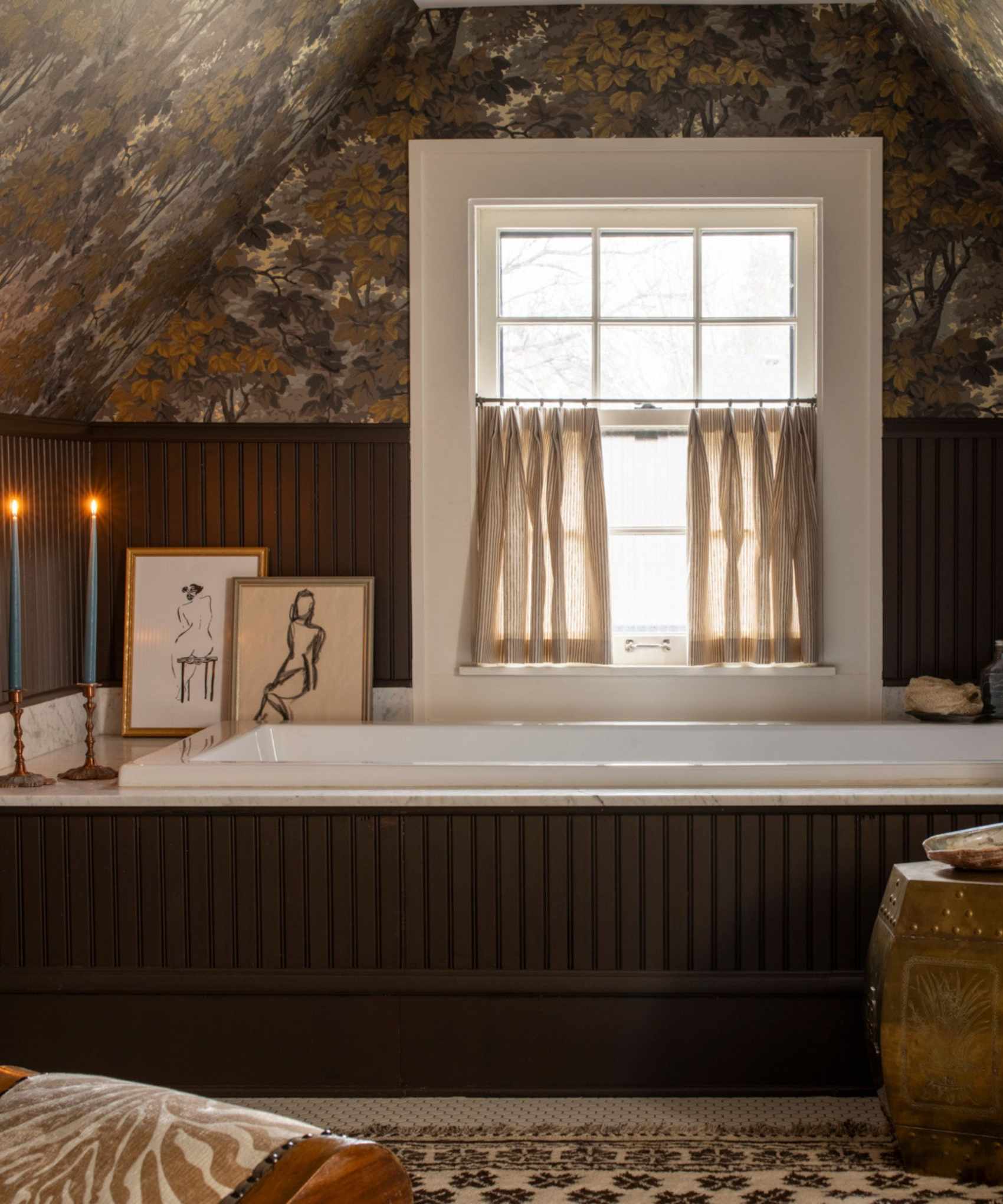 moody loft guest bathroom with dark wooden brown panelling and bath panel, small window with cafe curtain, botanical wallpaper, decorated with a rug, stool, artwork, candles and a small chair