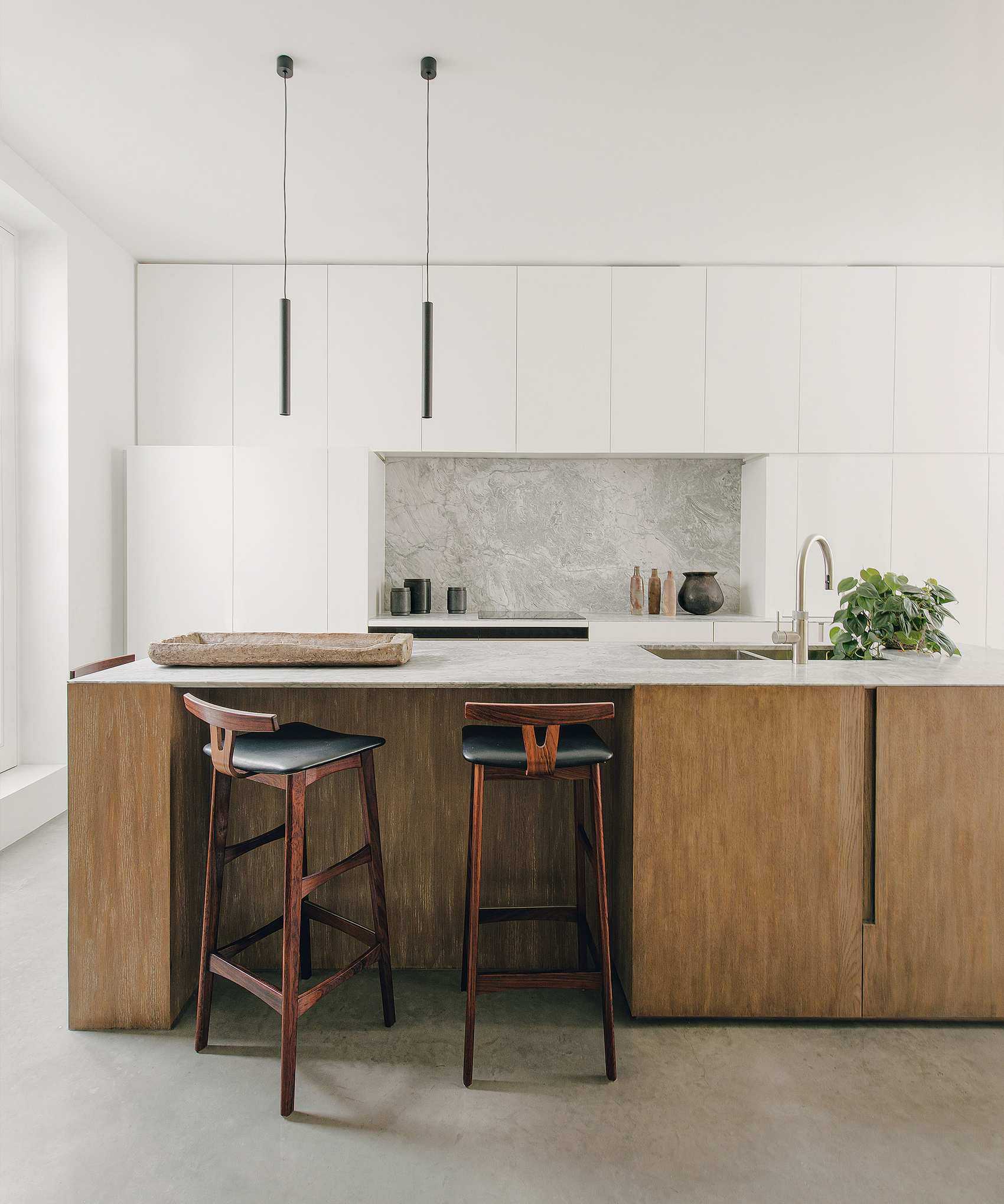 Modern kitchen space with kitchen island with dark wood base and cupboards, dark wood and black leather bar stools, white cupboards and white painted walls, two long black pendants hanging over island