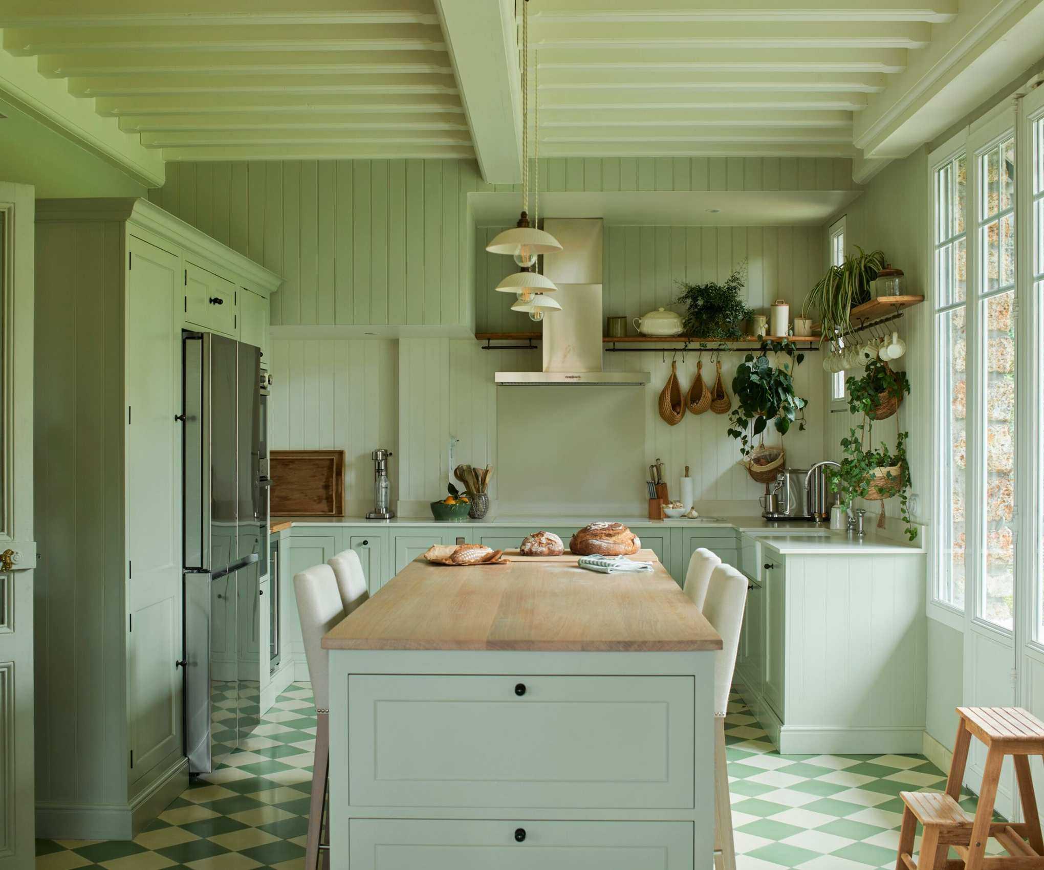 A pale green color-drenched kitchen with a wooden island countertop, a stainless steel range hood, and green and white checkerboard tiled flooring.